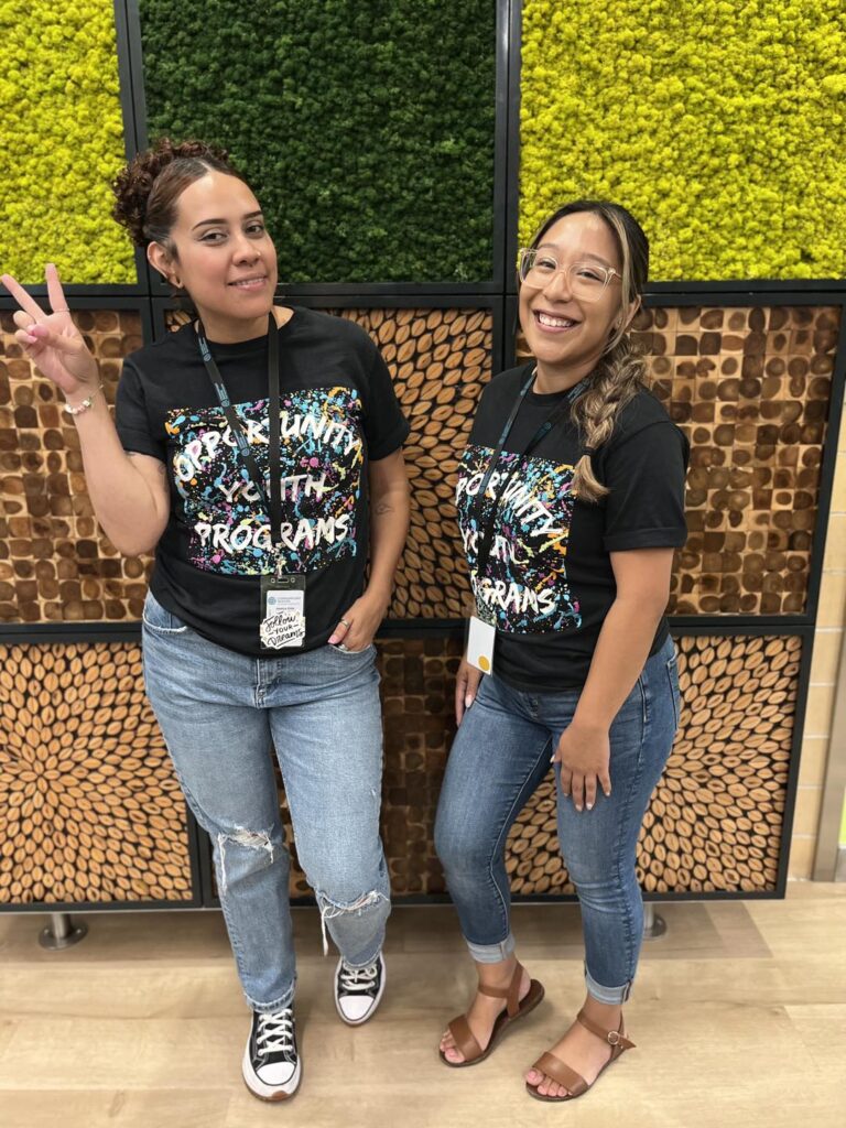 Two women in t-shirts participating in programs offered by the Youth Opportunity Hub, standing in front of a wall.