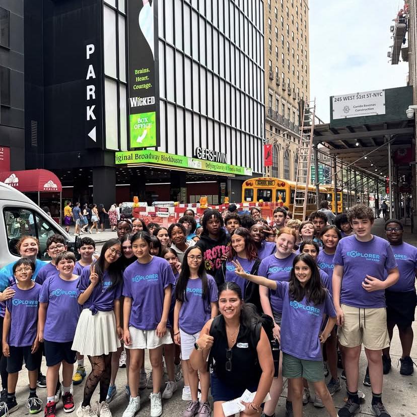 A large group of students and an adult, mostly wearing matching purple shirts, pose and smile for a photo on a busy city sidewalk in front of the Gershwin Theatre in New York City during their Explorer Day Camp outing.
