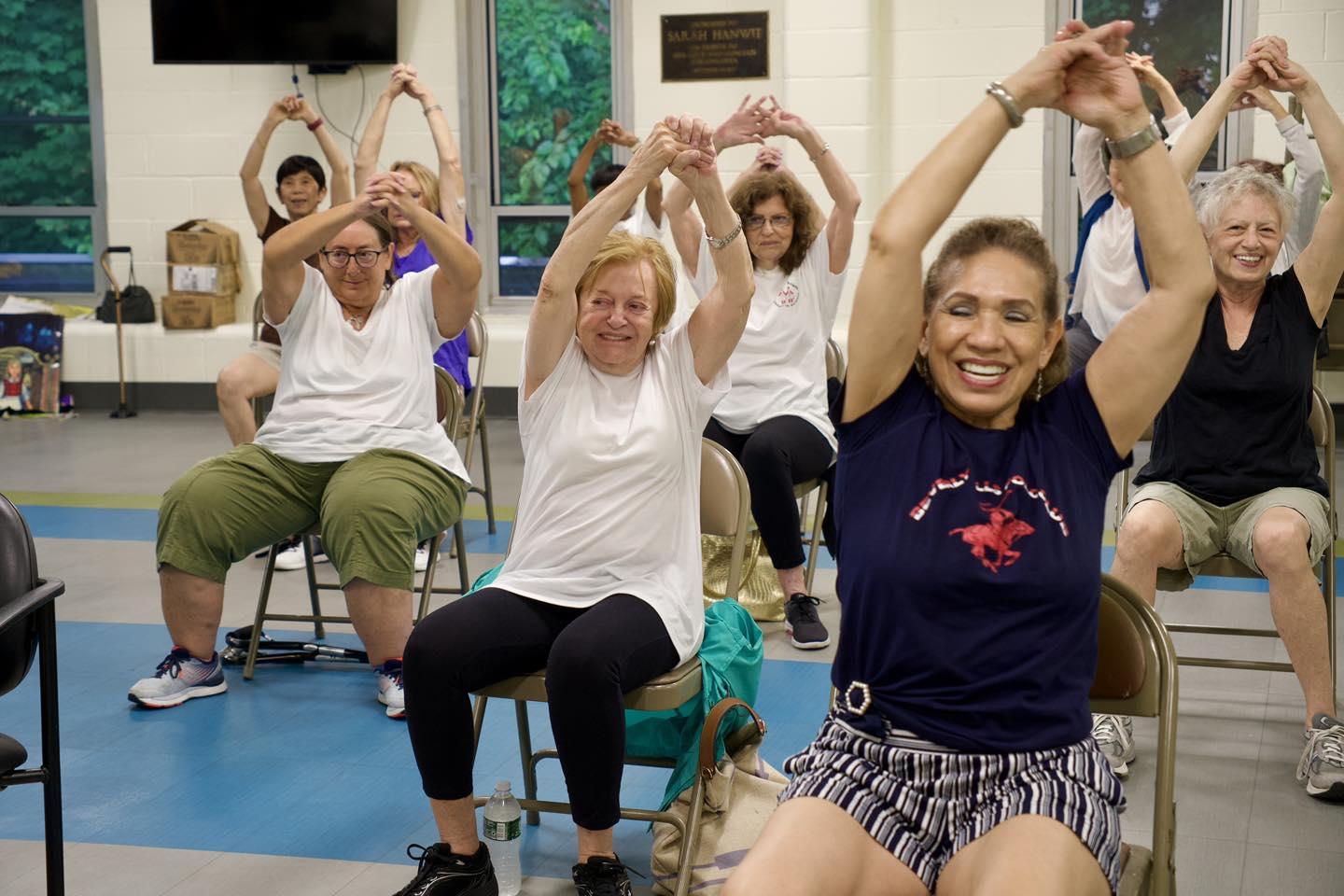 A group of older adults sit on chairs in the Sam Field gym, smiling and stretching their arms overhead during a fitness class at the Older Adult Center.