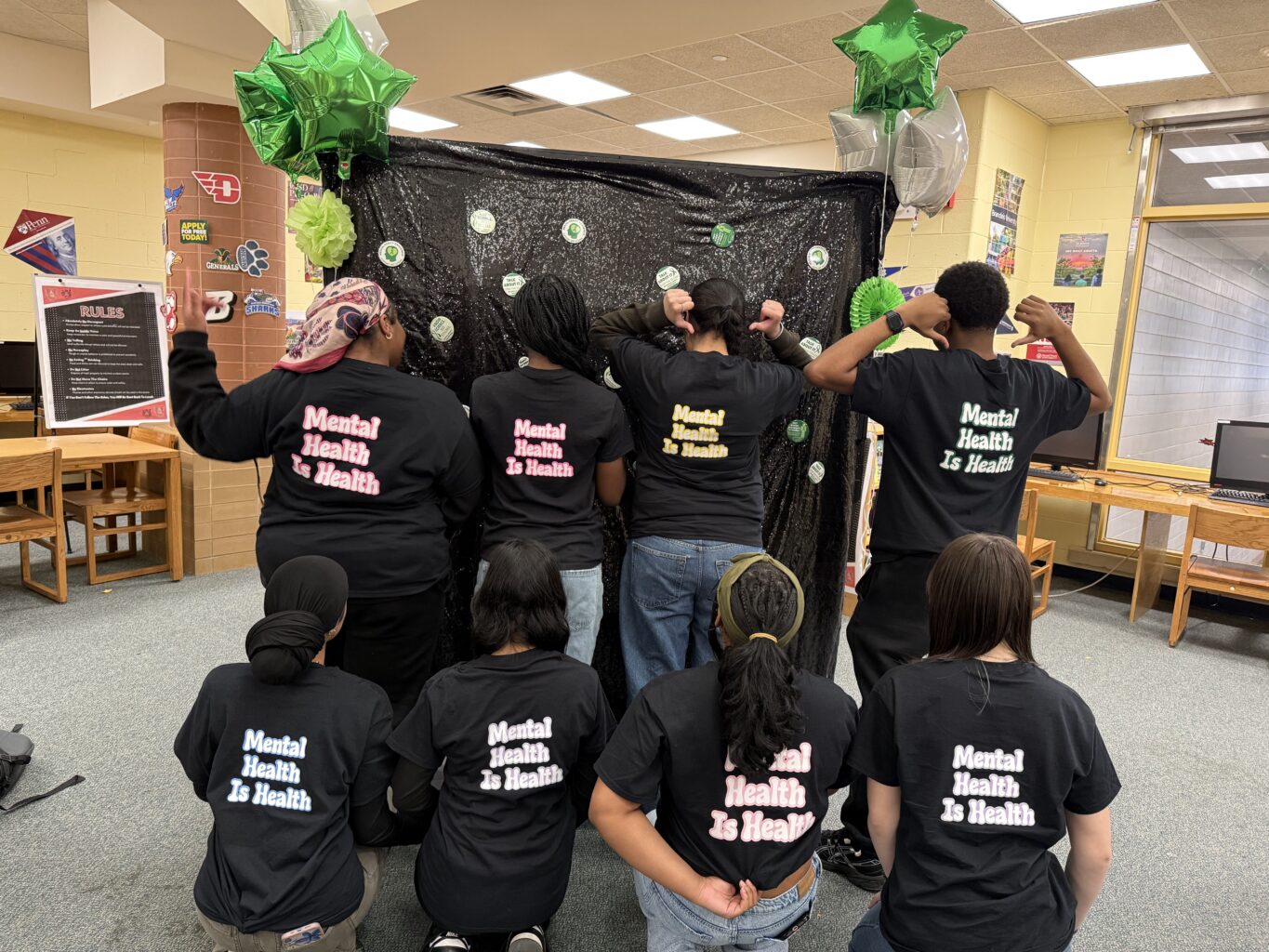 Nine people pose with their backs to the camera, wearing shirts that say “Mental Health Is Health,” in a decorated indoor space with green balloons overhead—a vibrant display highlighting the importance of Youth Mental Health and accessible Mental Health Services.