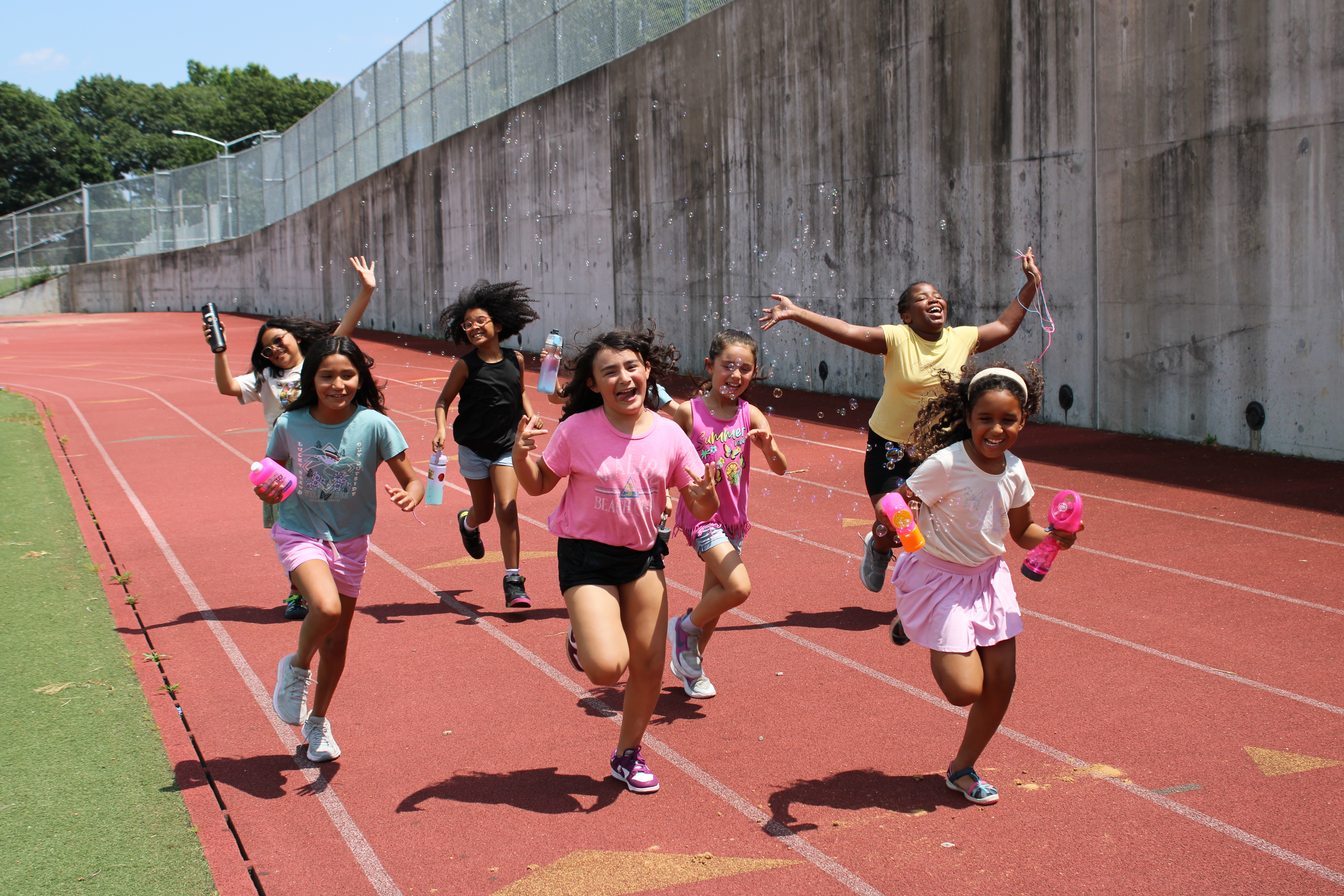 Seven girls run and laugh together on a red outdoor track at Explorer Day Camp, holding water bottles, with a concrete wall and chain-link fence in the background.