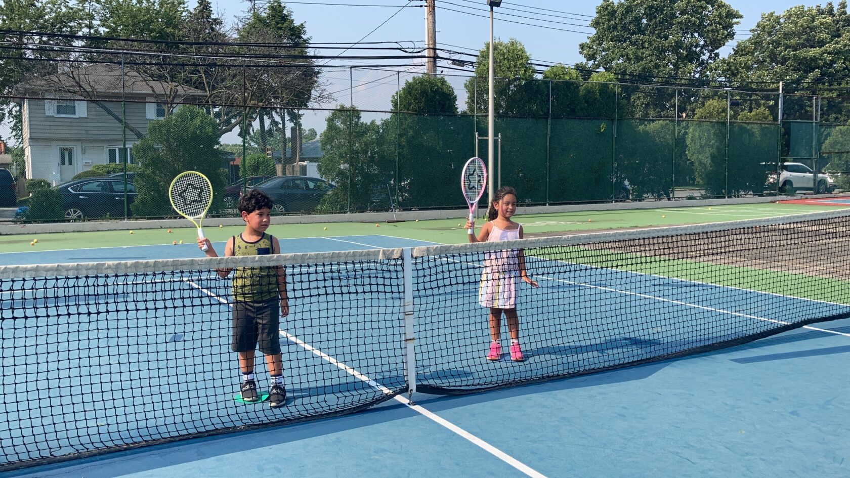 A young boy and girl stand on opposite sides of a tennis net holding plastic toy rackets during a youth tennis class.