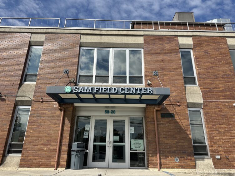 The entrance of the Sam Field Center features a brick building with large windows, a blue canopy, and a prominent Sam Field Center sign above the doors.
