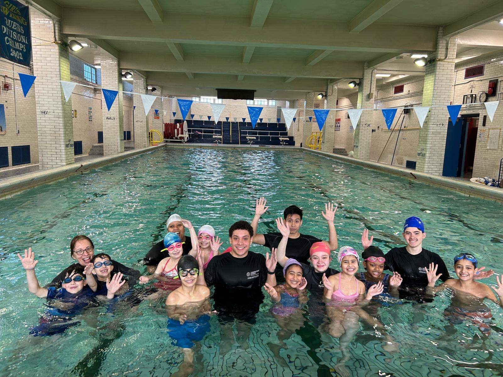 A group of children and adults in swim caps and goggles pose and wave in an indoor swimming pool during swim lessons, with bleachers and blue pennant banners in the background at Jamaica Hills.