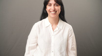 Albina Baazov, with long dark hair and a warm smile, stands in front of a plain gray background wearing a crisp white button-up shirt.