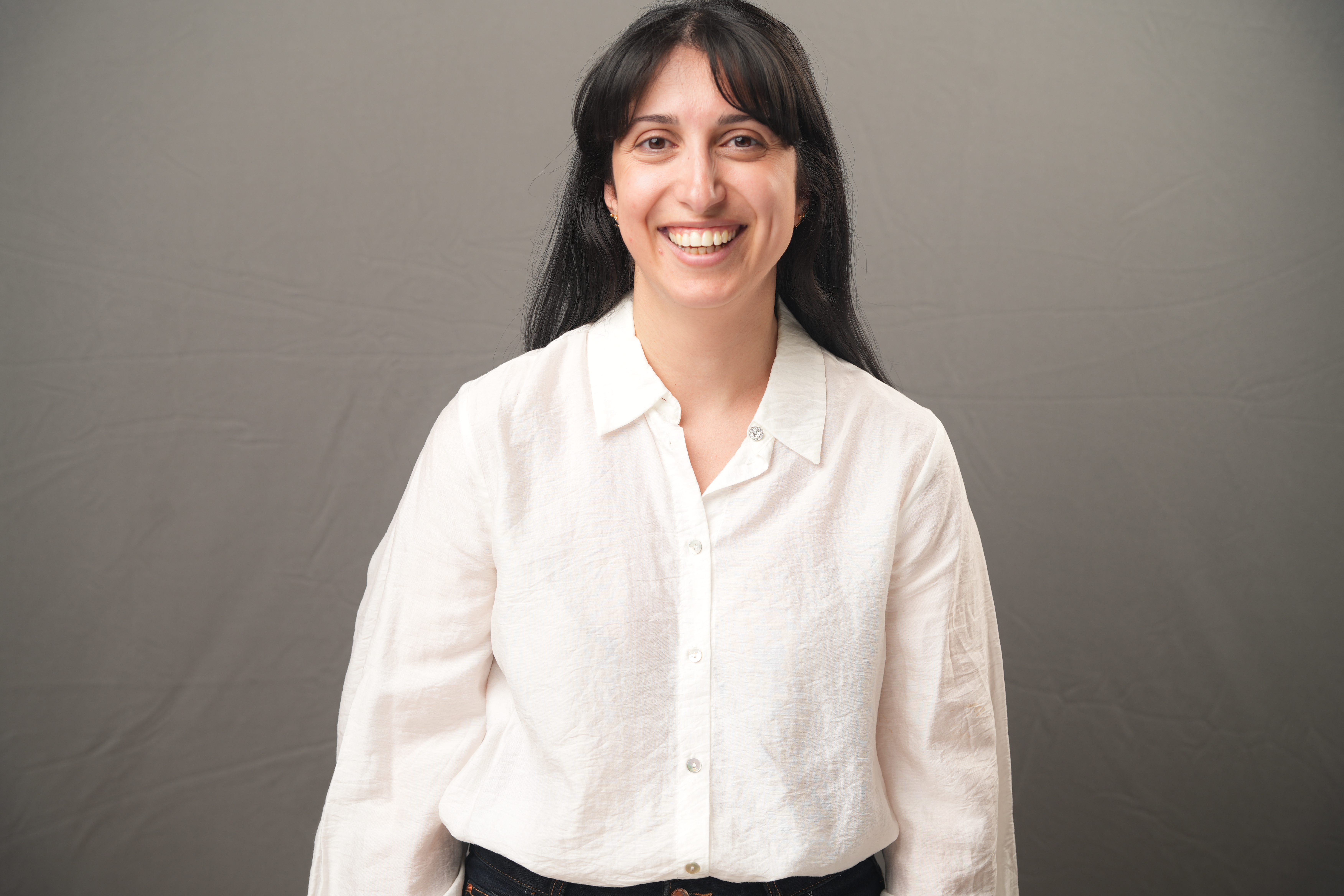 Albina Baazov, with long dark hair and a warm smile, stands in front of a plain gray background wearing a crisp white button-up shirt.