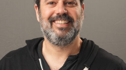 A man with a beard and long hair tied back, identified as Danny Mishkin, wears a black zip-up hoodie with "Sababa Beach" printed on it. He stands in front of a plain gray background, smiling at the camera.