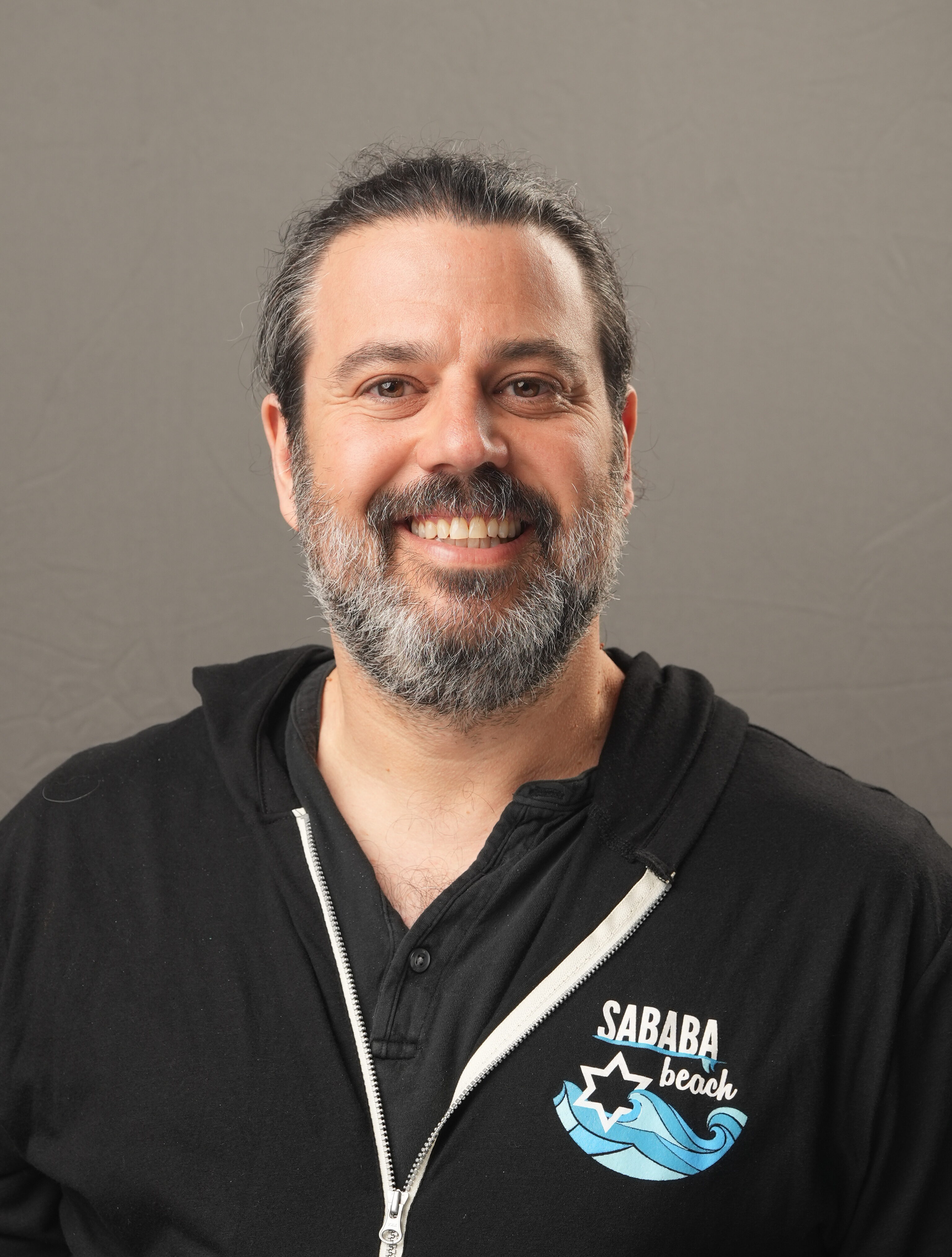 A man with a beard and long hair tied back, identified as Danny Mishkin, wears a black zip-up hoodie with "Sababa Beach" printed on it. He stands in front of a plain gray background, smiling at the camera.