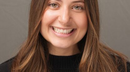A woman with long brown hair and a black sweater smiles in front of a plain gray background.
