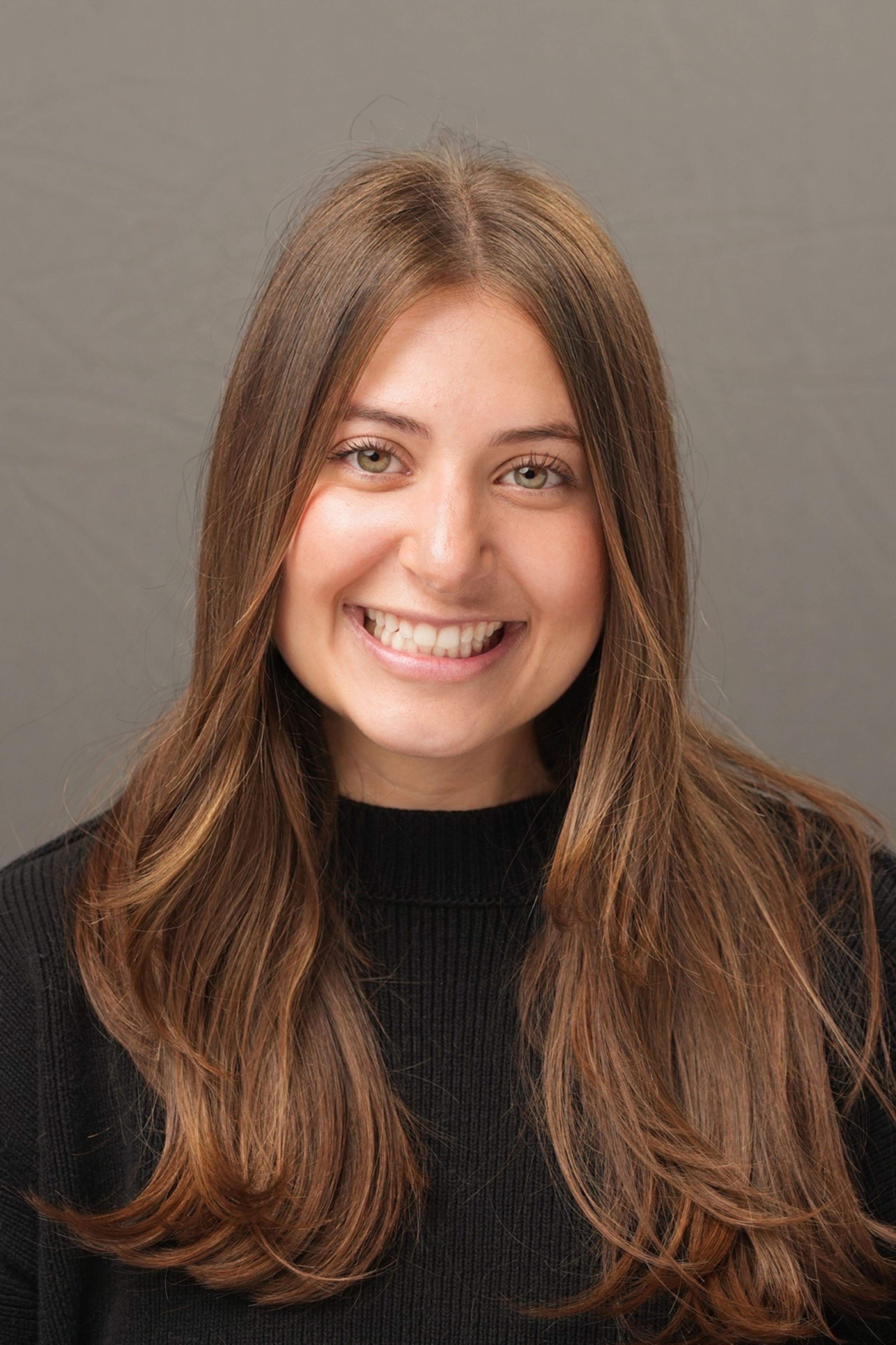 A woman with long brown hair and a black sweater smiles in front of a plain gray background.