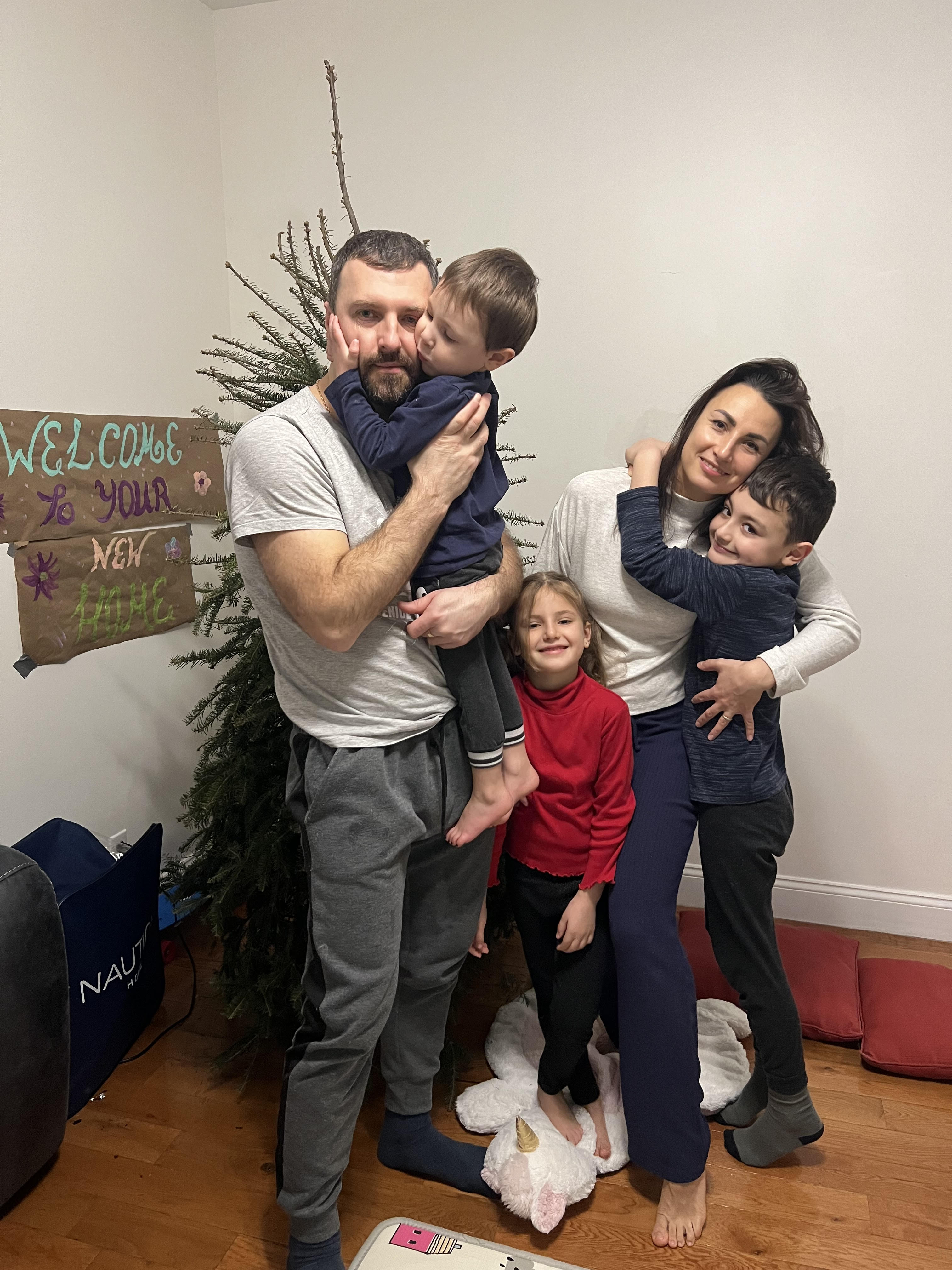 A family of two adults and three children stand in front of a decorated tree, with a “Welcome to your new home” sign, celebrating their resettlement. The parents hold two children in their arms while the third child stands in front.