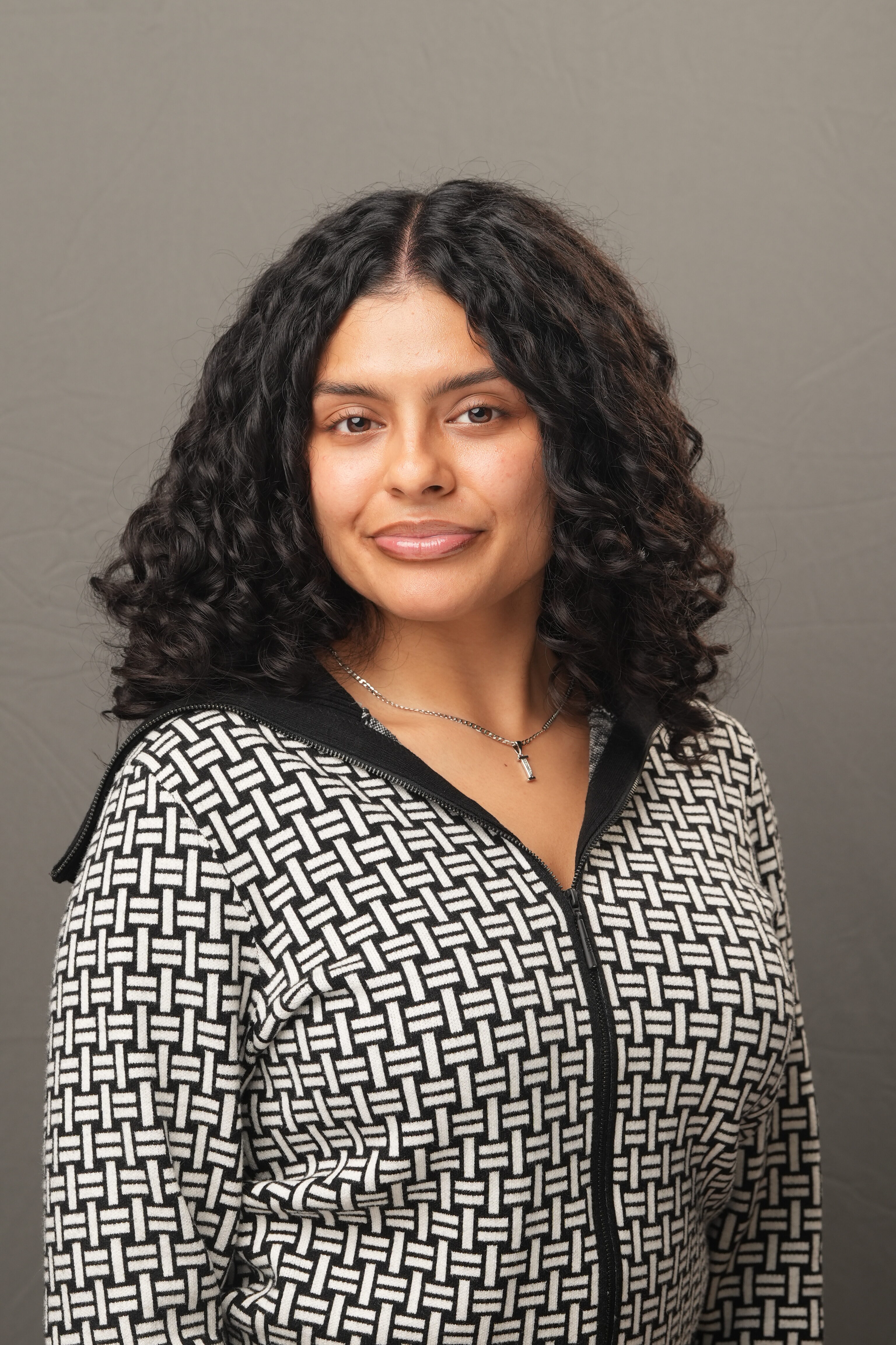 A woman with curly dark hair wearing a patterned zip-up sweater and a necklace stands in front of a plain gray background, facing the camera.