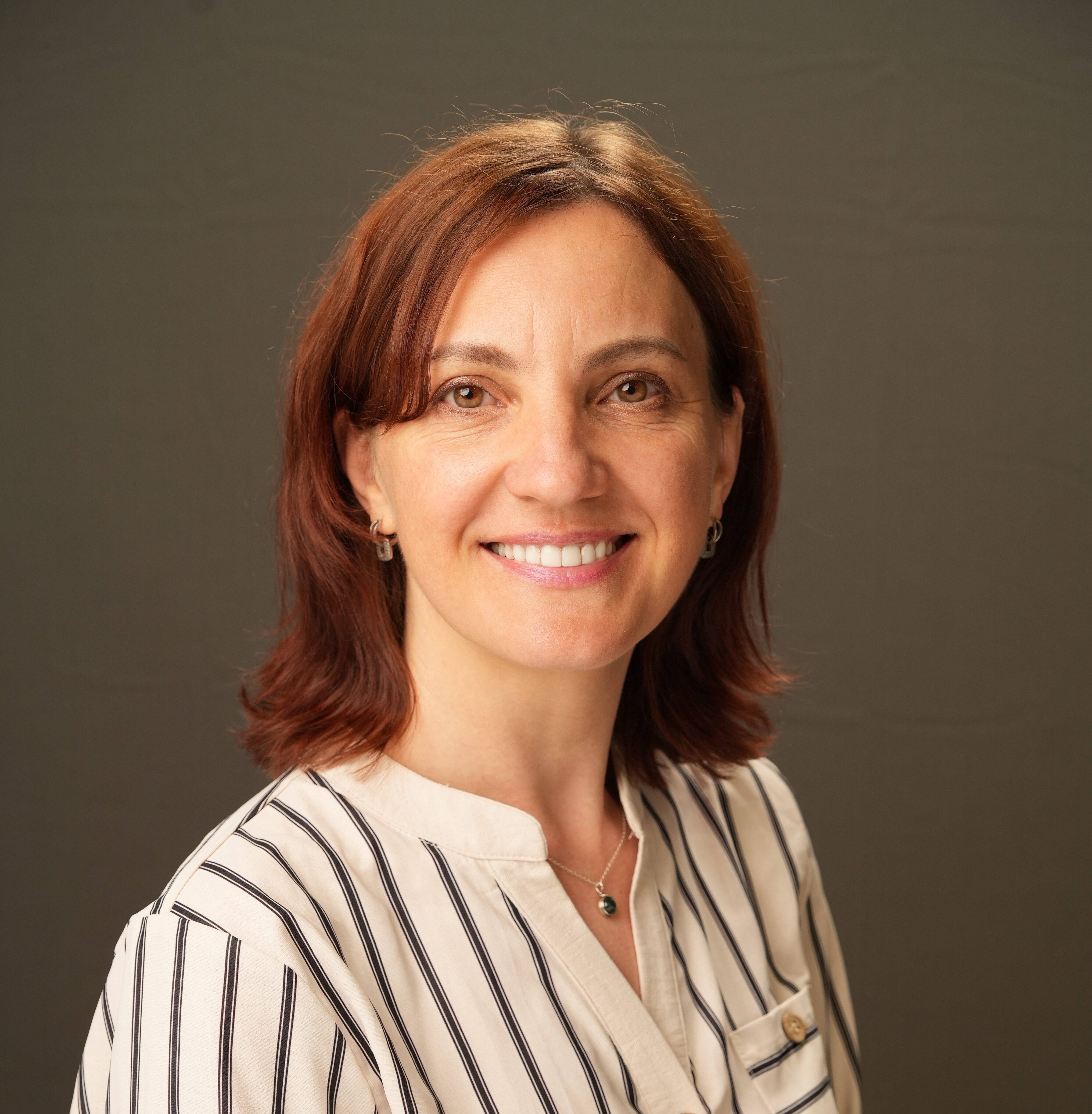 Oksana Lysyshyn, a woman with shoulder-length brown hair, wearing a white shirt with black vertical stripes, stands smiling in front of a plain gray background.