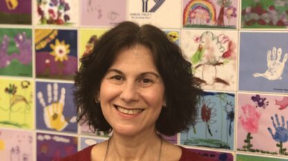 Robin Kaufman, a woman with dark hair in a red sweater, stands smiling in front of a colorful wall display featuring children's handprints and artwork.