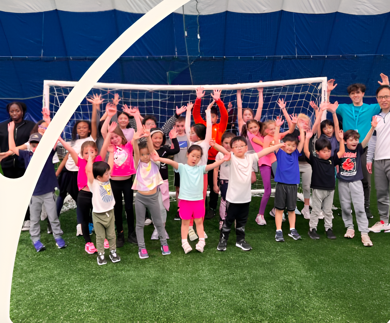 A group of children and adults stand together, raising their hands in front of a soccer goal inside a sports dome, embodying the spirit of Camp 2 at Commonpoint Presents.