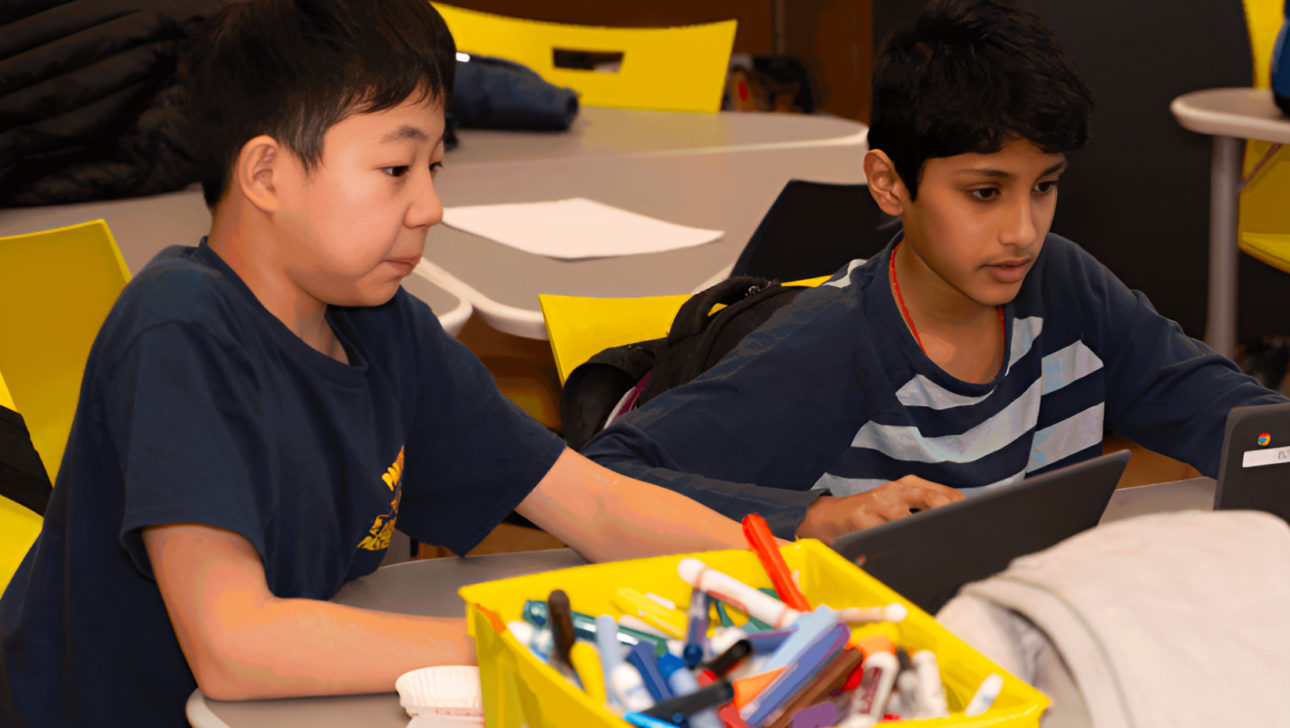 Two children sitting at a table using tablets after school, with a yellow container filled with markers and stationery in the foreground. They appear focused on their screens.