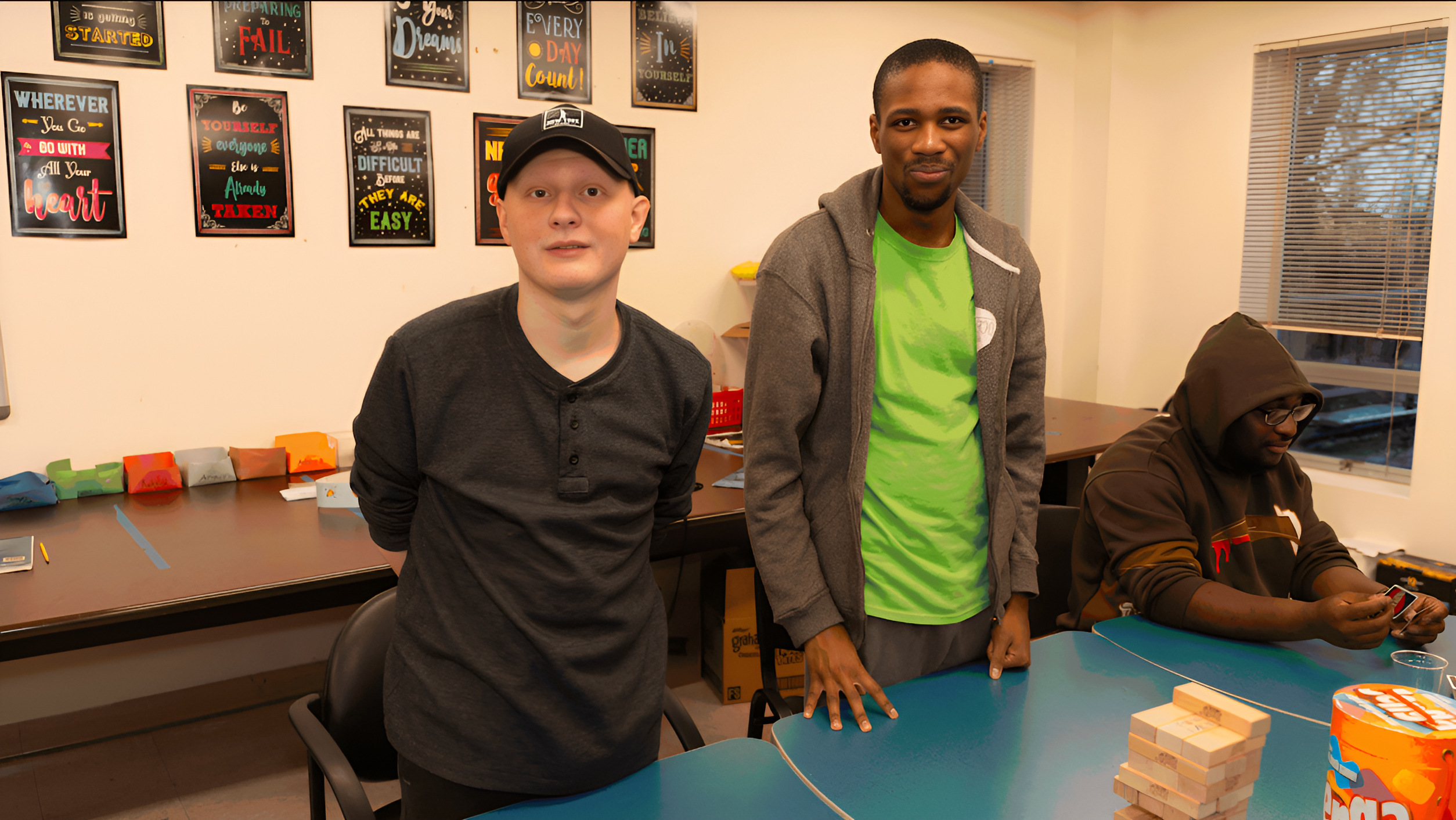 Three people are in a room by a table with a Jenga game. One person stands on the left, another in the center, and the third person is sitting on the right using a phone. Decor and games from Commonpoint's special services are in the background.