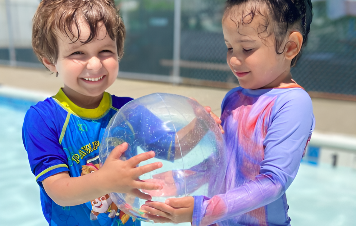 Two children in swimwear hold a beach ball while standing in a shallow pool at Camp 2 Commonpoint. Both are smiling and appear to be having fun under the sun.