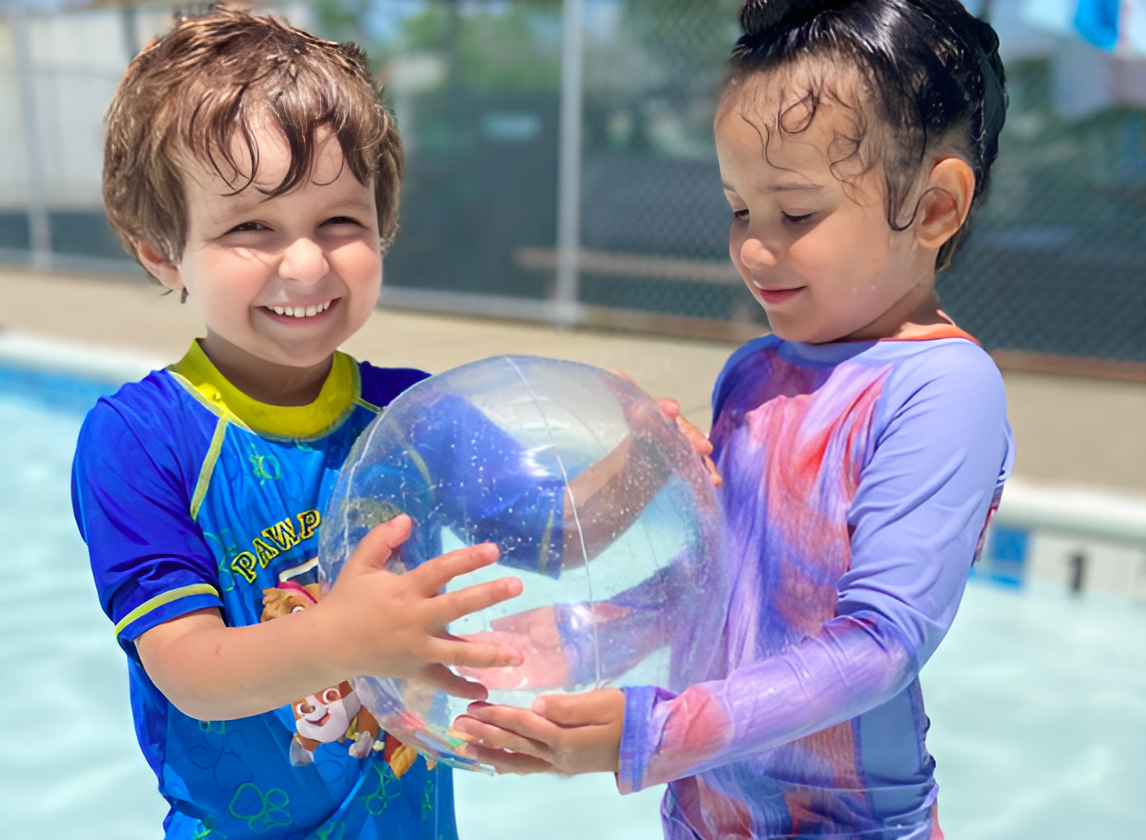 Two children in swimwear hold a beach ball while standing in a shallow pool at Camp 2 Commonpoint. Both are smiling and appear to be having fun under the sun.