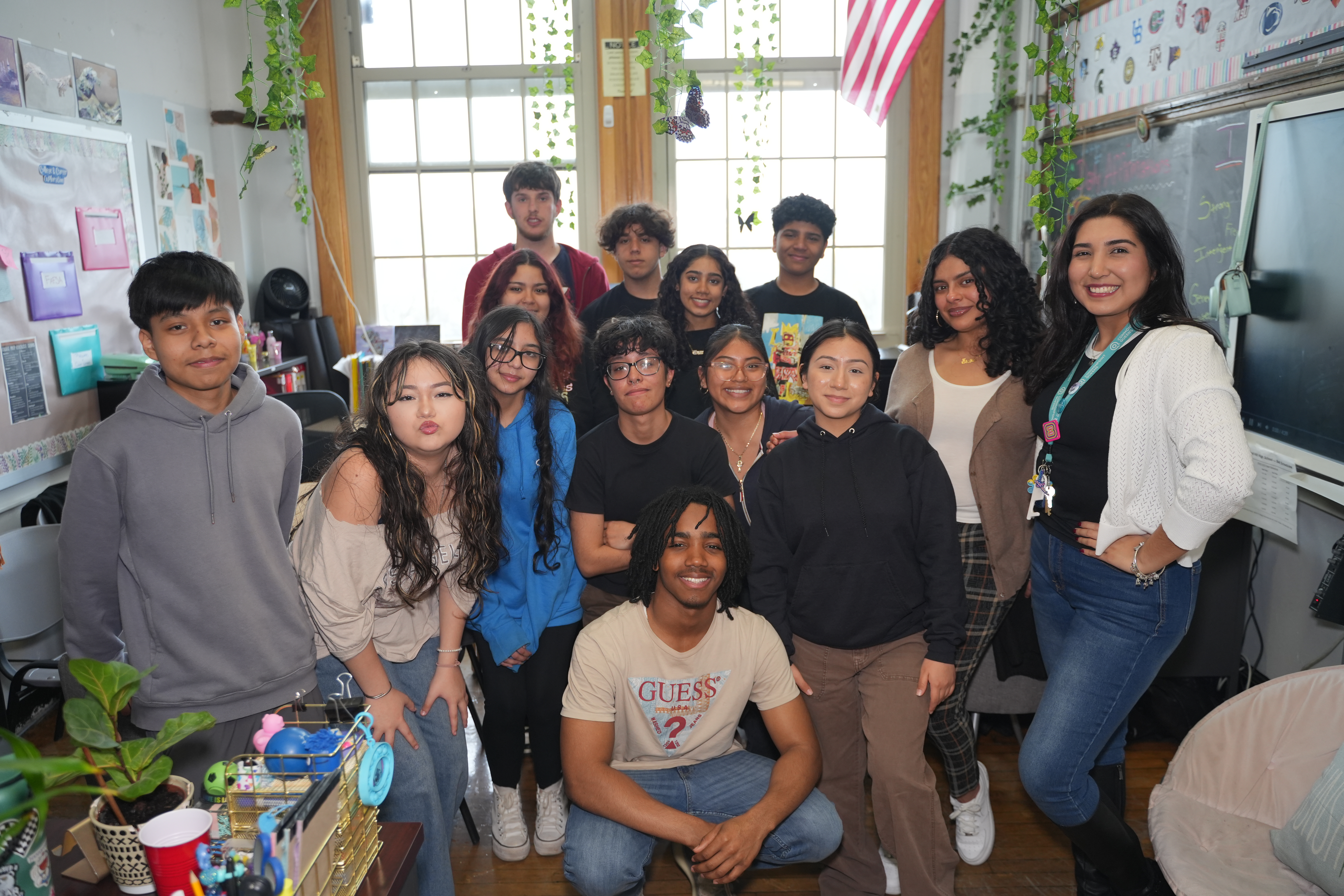 A group of students and a teacher pose together in a classroom with plants and an American flag in the background, capturing the spirit of their DYCD Hip-Hop Holiday Celebration.
