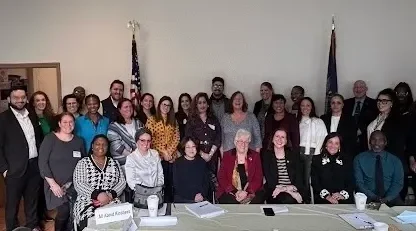 A diverse group of people pose together indoors in a meeting room, discussing culturally competent services. A table with paperwork and nameplates sits in the foreground while two flags are displayed in the background, emphasizing their commitment to inclusive solutions.