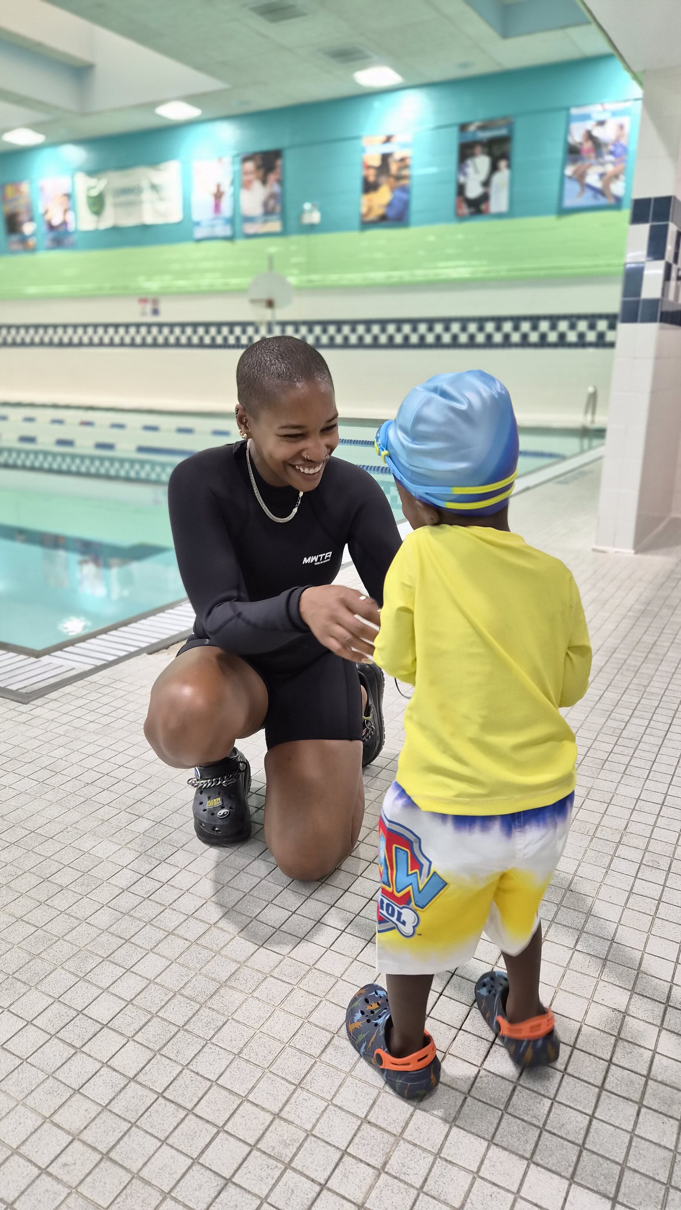 An adult in a black swimsuit kneels and smiles at a young child in swim gear on the deck of an indoor swimming pool, capturing a heartwarming moment during Bronx Aquatics swim classes.