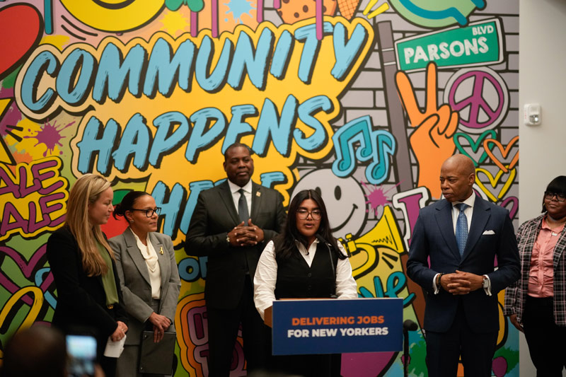 A group of six people stands in front of a colorful mural with the words "Community Happens Here." A podium reads "Delivering Jobs for New Yorkers," highlighting Mayor Adams' $15 million investment to boost job opportunities across the city.