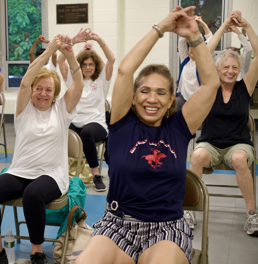 In a lively gym, a group of people, all smiles, stretch their arms overhead as they participate in an Older Adult Services fitness class. Dressed in casual workout gear, they enjoy the vibrant atmosphere and focus on wellness together.