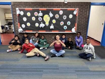 A group of children and an adult sit on the floor in front of a decorated bulletin board labeled "Commonpoint Bronx." Some kids make heart and peace signs with their hands, adding a warm touch to the school hallway. It feels like a moment from Summer Camp.