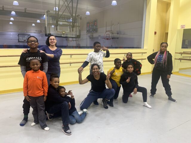 A group of children and two adults pose together in a dance studio at Commonpoint Bronx, with mirrors, ballet barres, and a gym visible in the background.