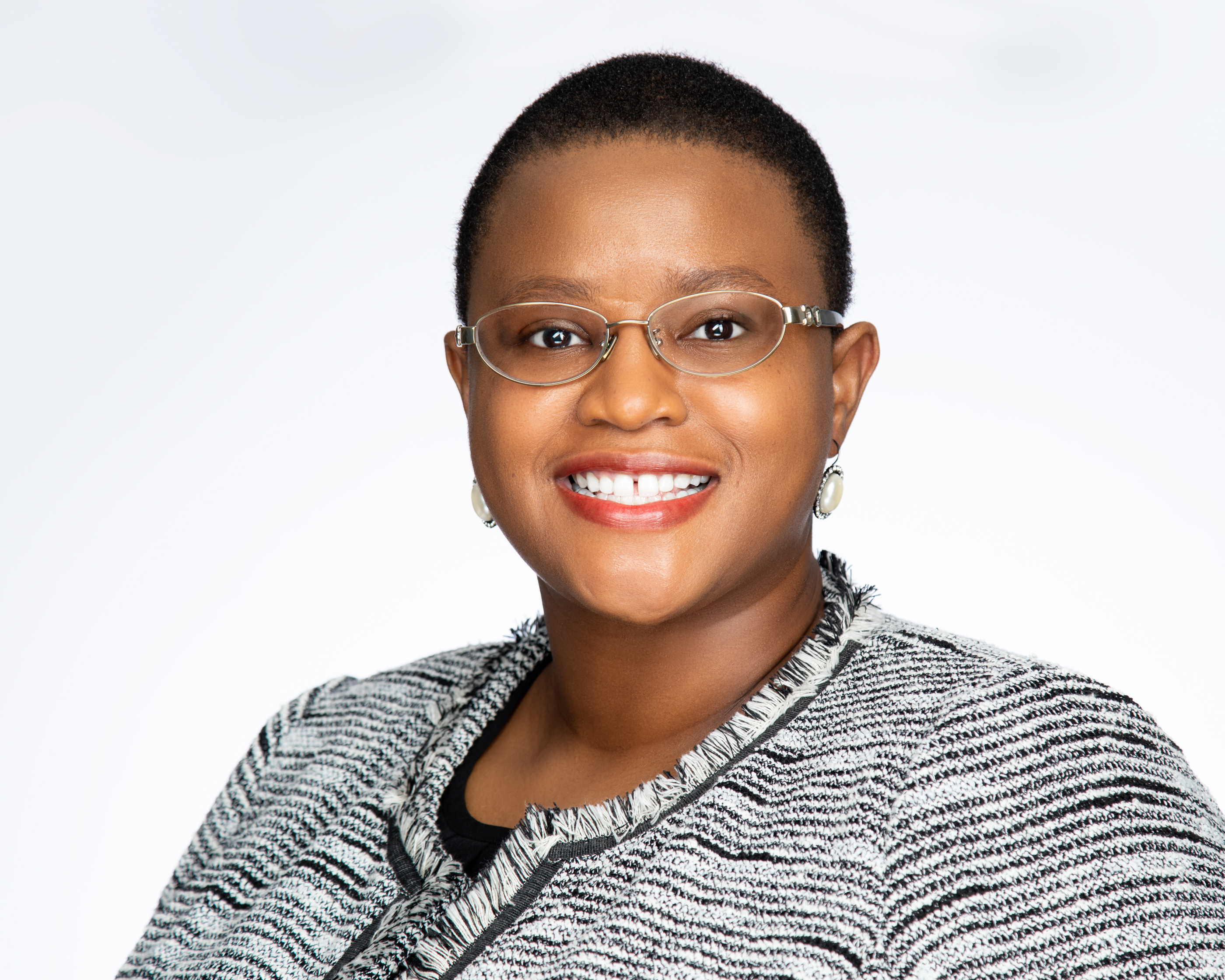 A beaming Kelenda Allen-James, sporting short hair and stylish glasses, stands confidently in a textured jacket against a plain background.