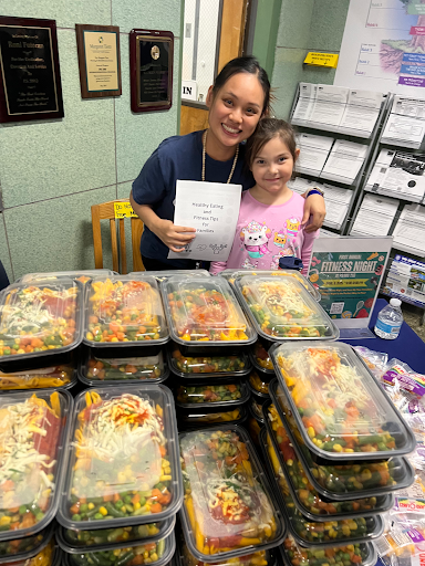 A woman and a child stand behind a table with stacked food containers filled with colorful vegetables. The woman holds a paper with "Healthy Eating Program" printed on it. Plaques hang on the wall.