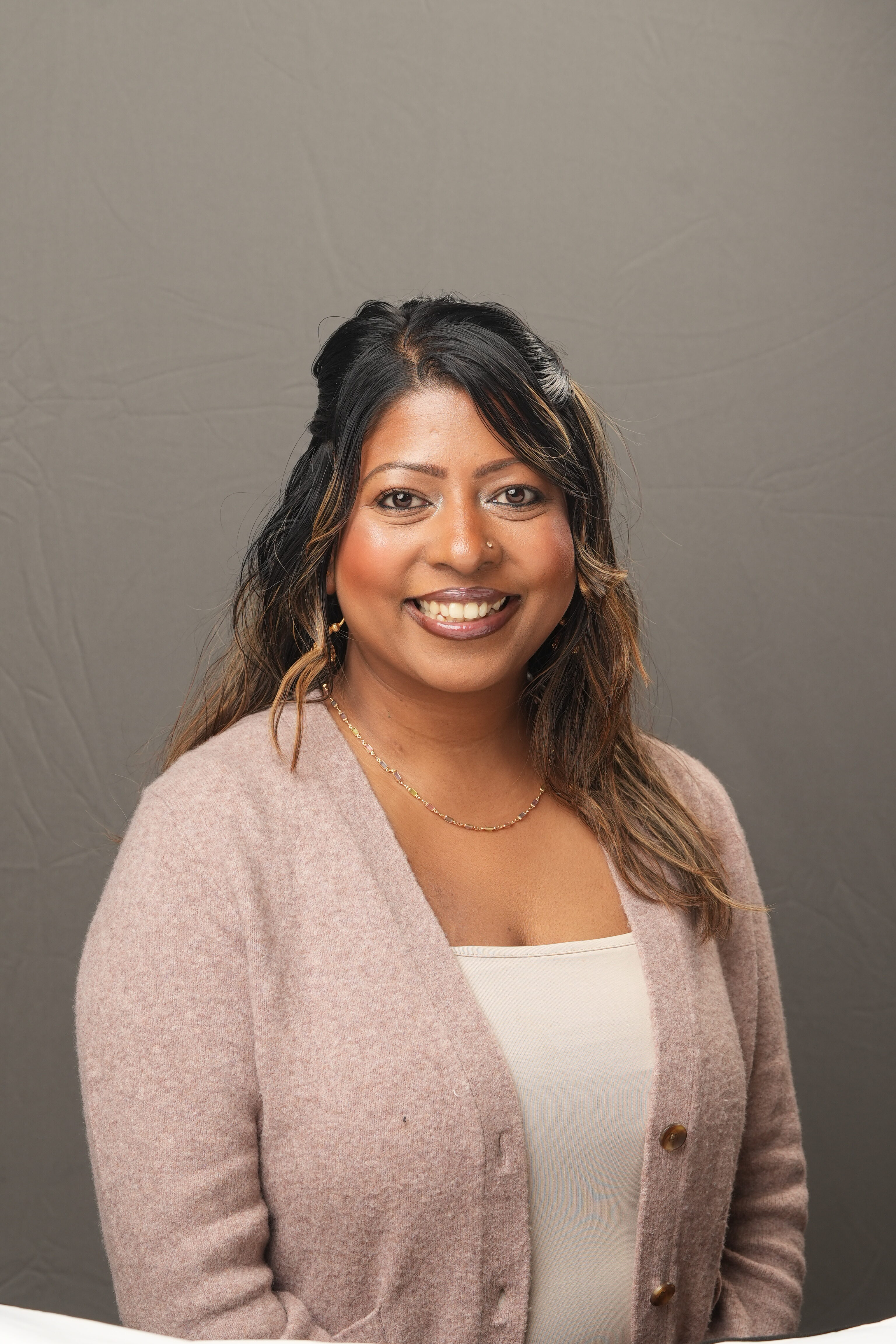 Serila Saimon, a woman with long dark hair wearing a light pink cardigan and cream top, is smiling at the camera against a plain gray background.