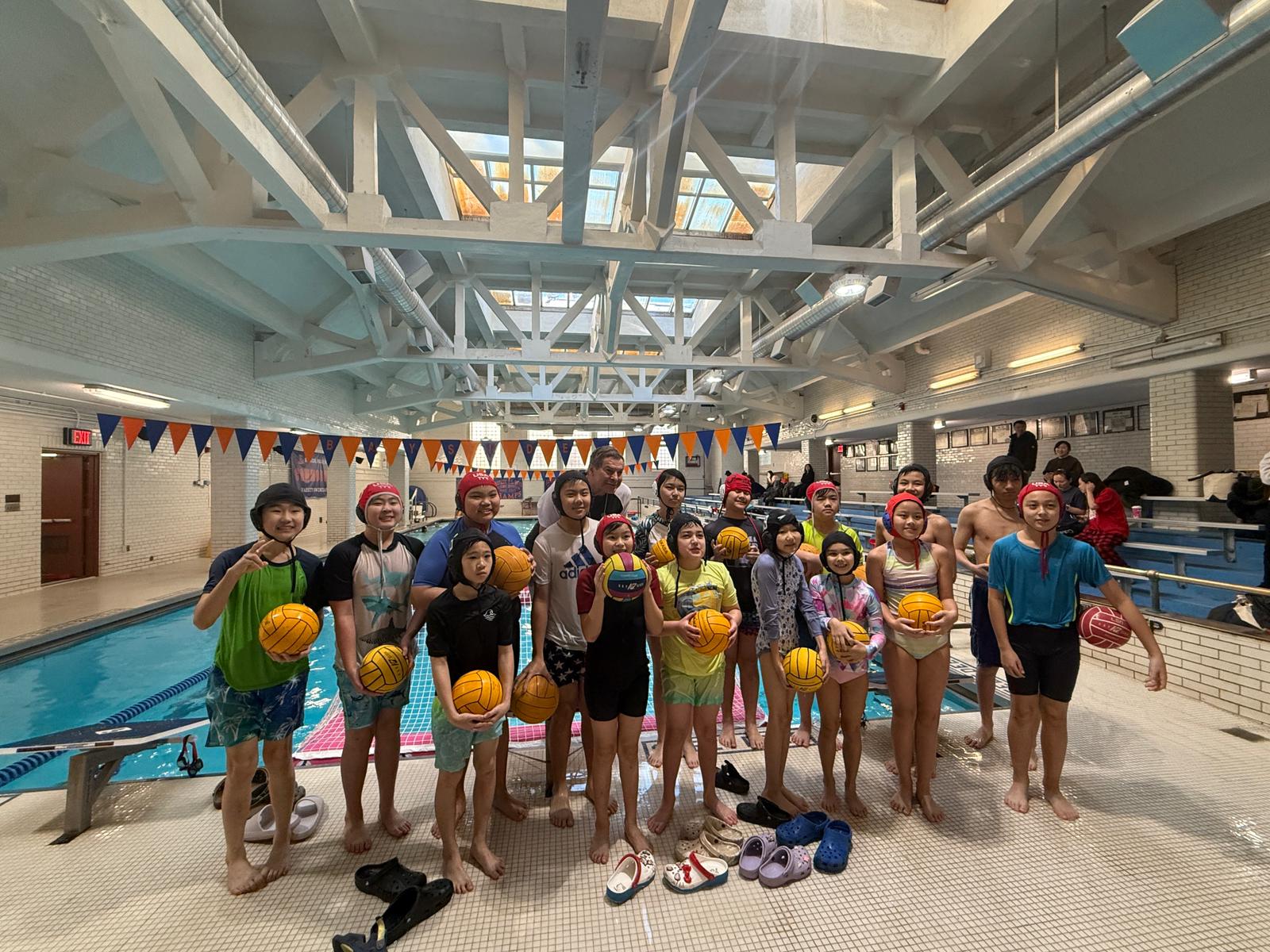 A group of children and adults in swimsuits stand beside an indoor pool at Bayside Water Polo Club, each holding a water polo ball, with orange and blue pennant banners hanging above.