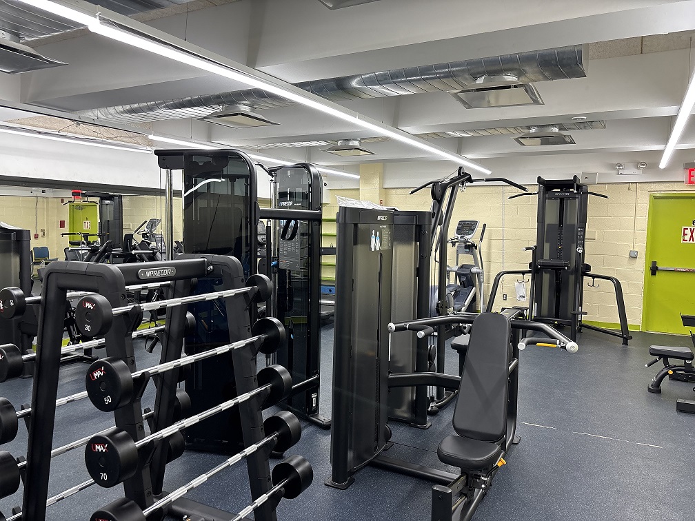 A gym with weight racks, various strength training machines, and cardio equipment, set in a brightly lit room with yellow walls and a green exit door—perfect for those with a Commonpoint Bronx Membership.
