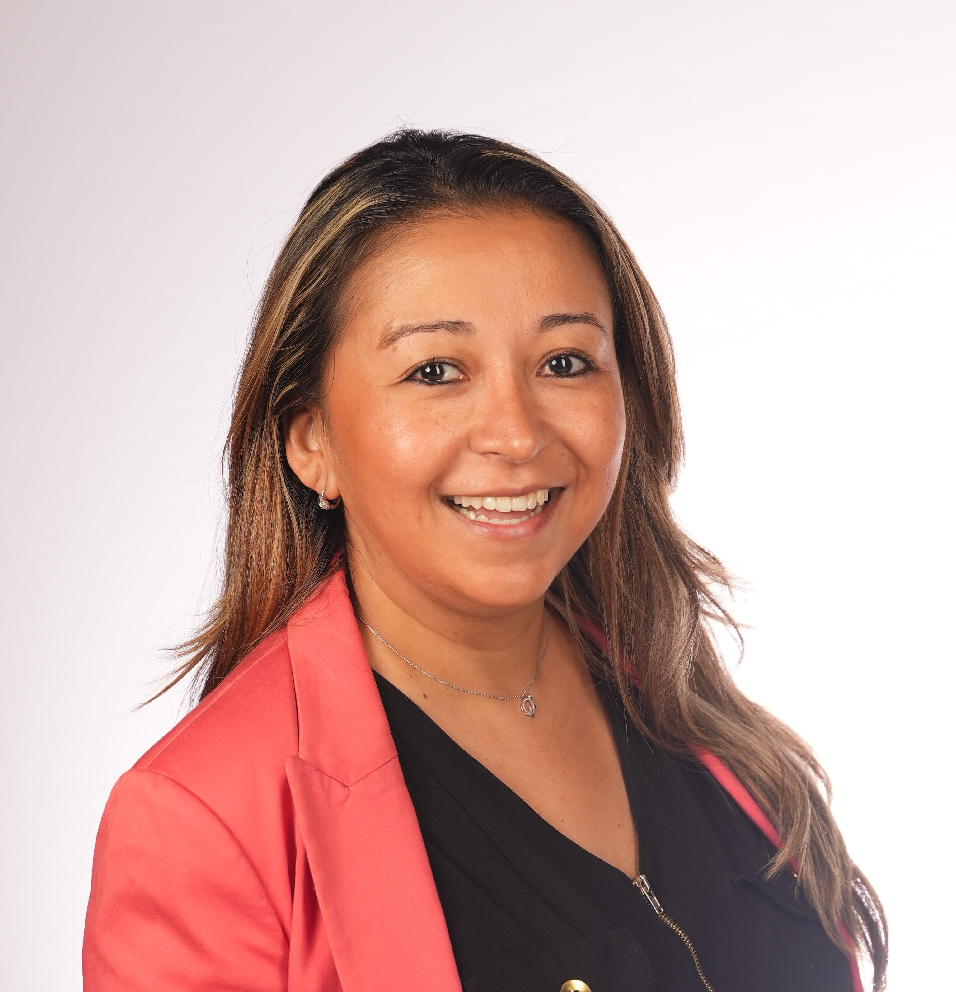 Nathalia Velasquez, a woman with long brown hair, smiles in a coral blazer over a black top against a plain light background.