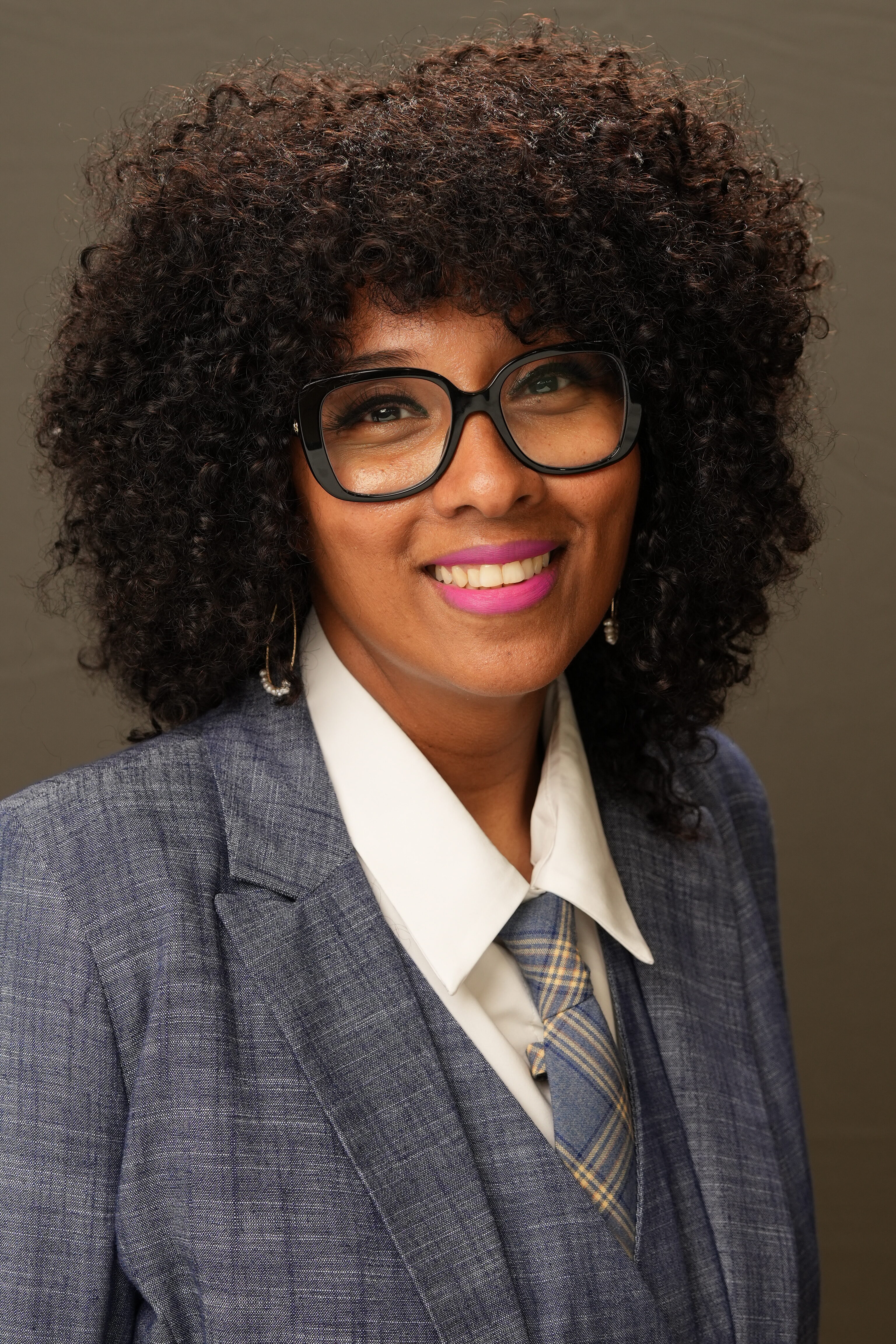 A person with curly hair, wearing glasses and a blue plaid suit with a plaid tie, smiles at the camera against a neutral background.