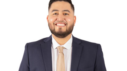 A man wearing a navy blue suit, light dress shirt, and beige tie stands facing forward with his hands in his pockets, smiling against a white background.