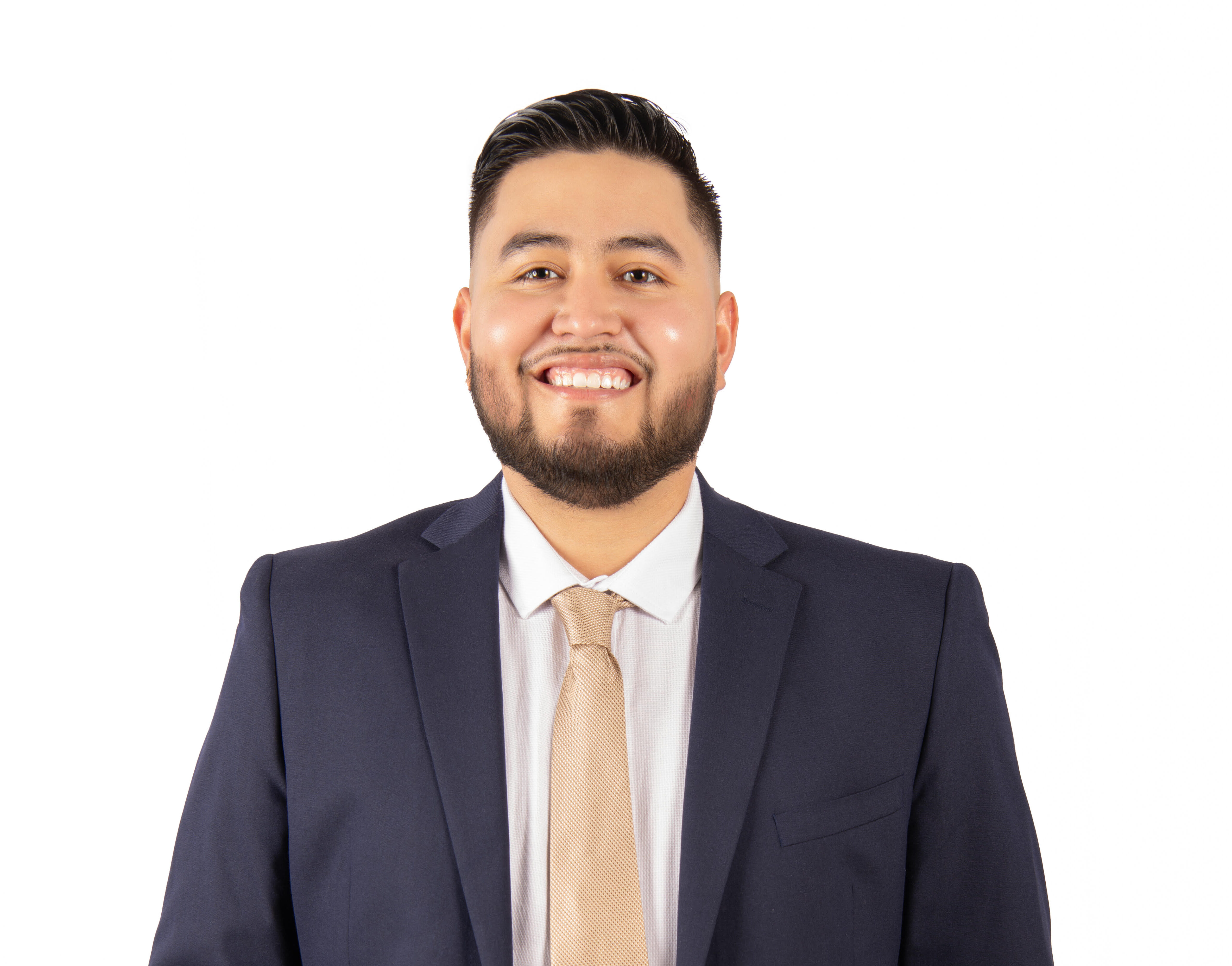 A man wearing a navy blue suit, light dress shirt, and beige tie stands facing forward with his hands in his pockets, smiling against a white background.