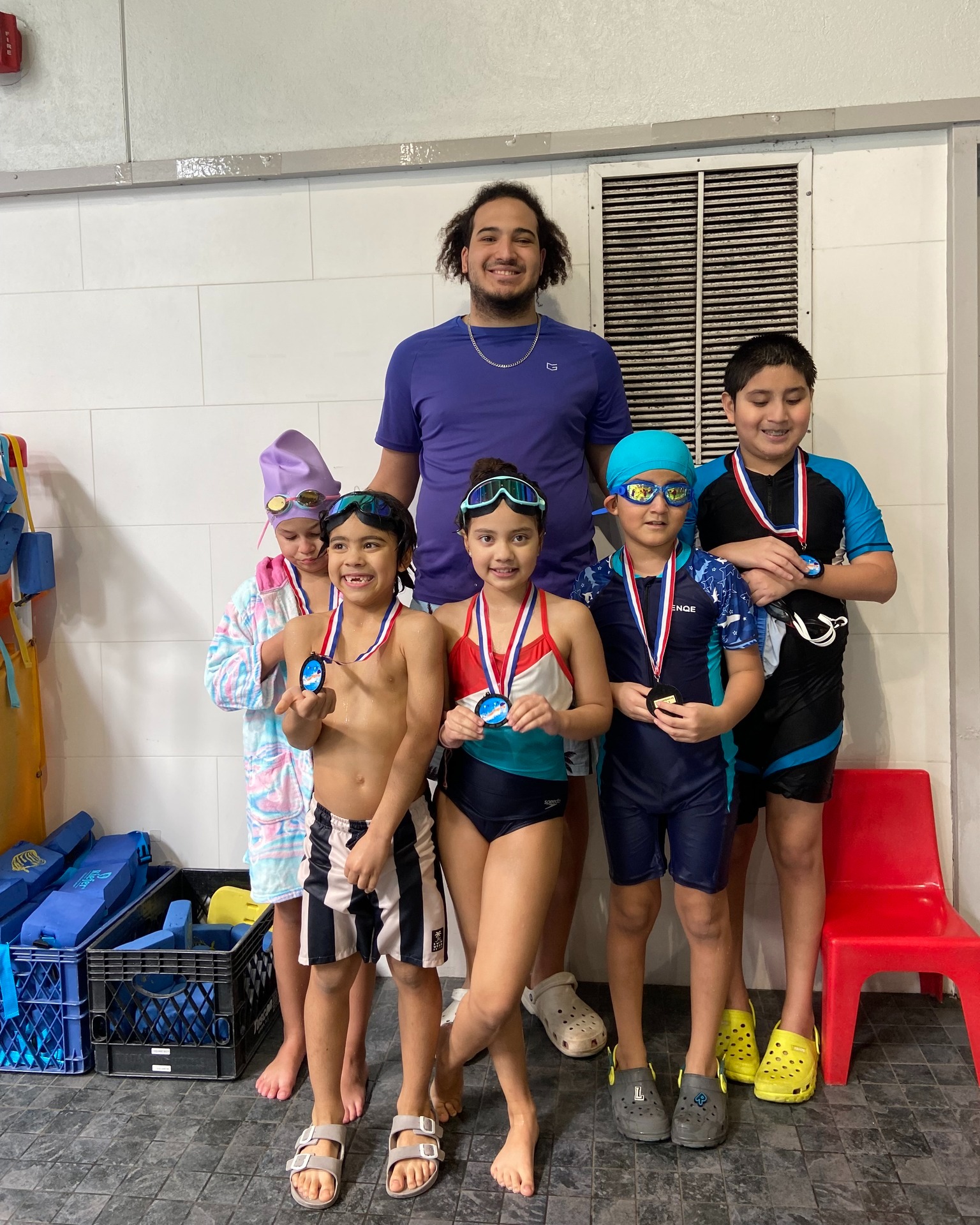 A group of five children wearing swim gear and medals stand with an adult in front of a tiled wall at an indoor pool, celebrating their achievements from the Fast Trac Swim Program.