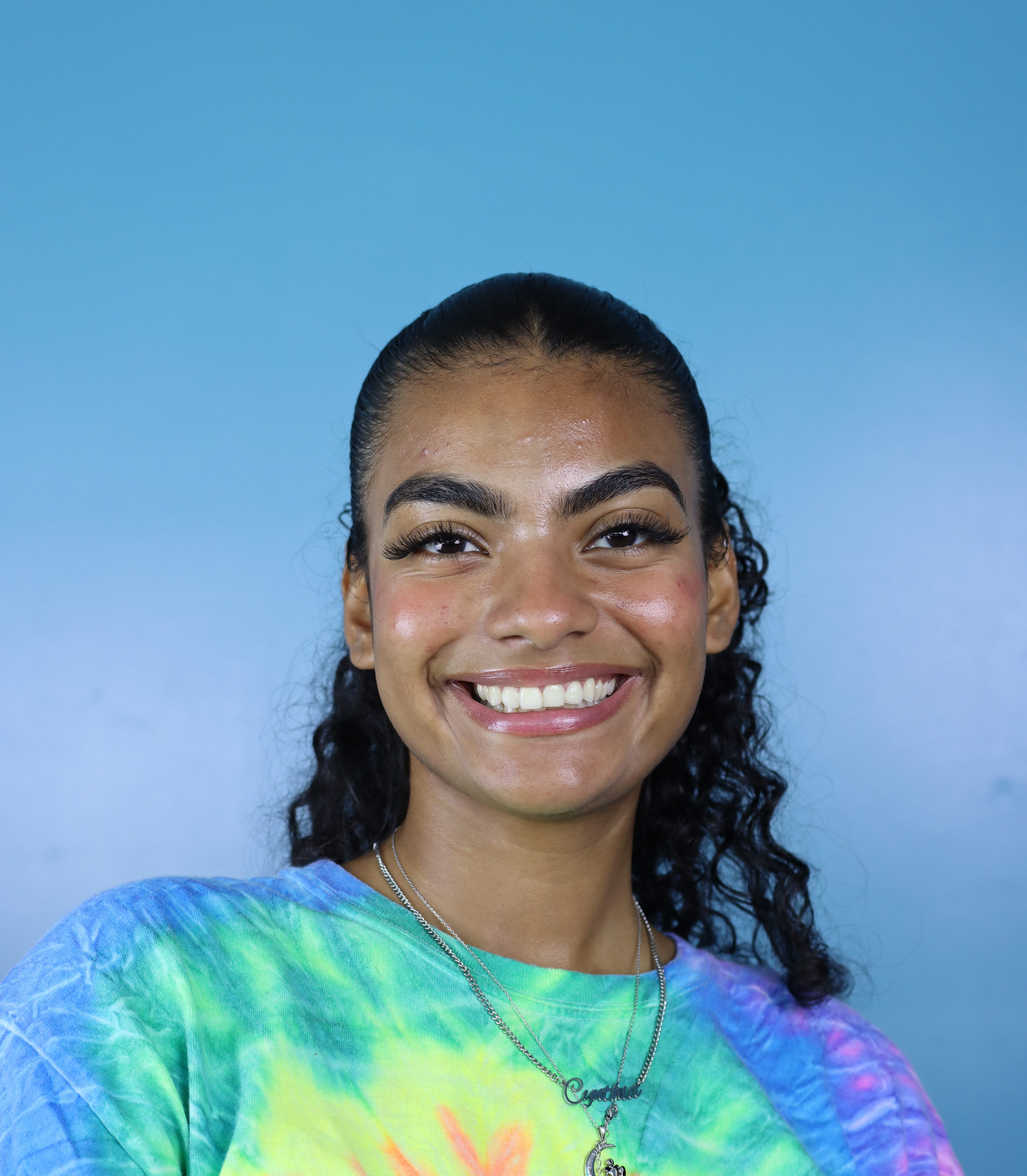 Cynthia Guzman, with curly hair, smiles at the camera in a colorful tie-dye Commonpoint Queens shirt, set against a vibrant blue background.