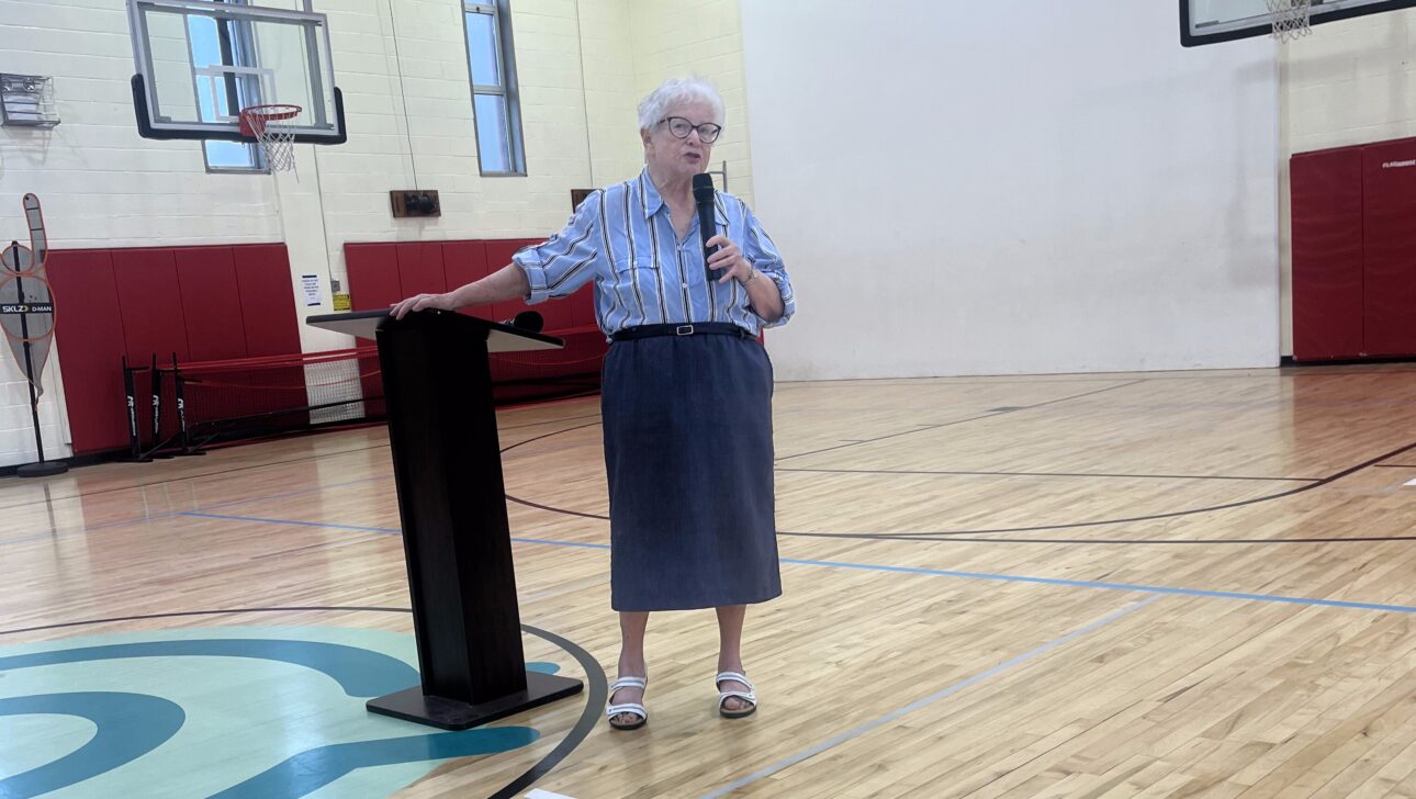 An older woman stands at a podium, speaking into a microphone in a gymnasium with basketball hoops and wooden floors during a Model Film Screening on Aging in Community in New York.