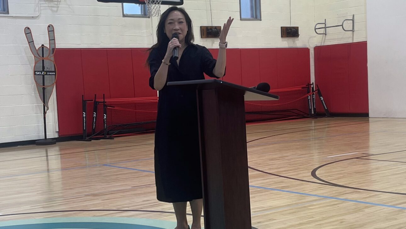A woman speaks into a microphone while standing at a podium in a gymnasium with red padding and basketball hoops during an Aging in Community event in New York.