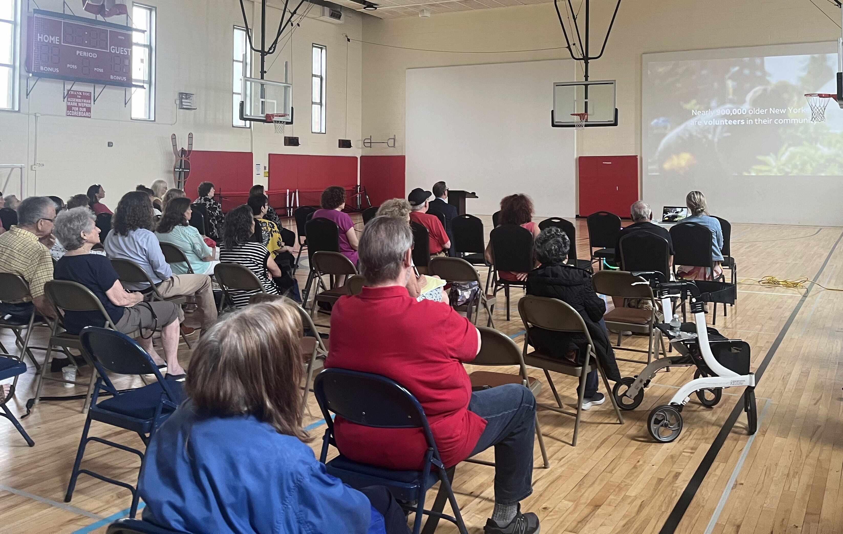 A group of people seated in a gymnasium in New York watch an Aging in Community film screening projected onto a screen at the front of the room.