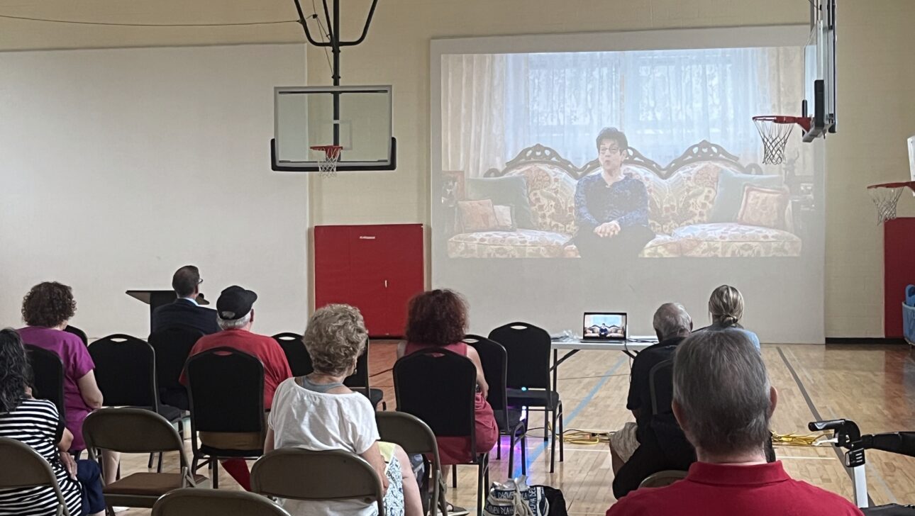 People sit on chairs in a gymnasium in New York, watching a film screening about Aging in Community, projected onto a screen set up in front of basketball hoops.