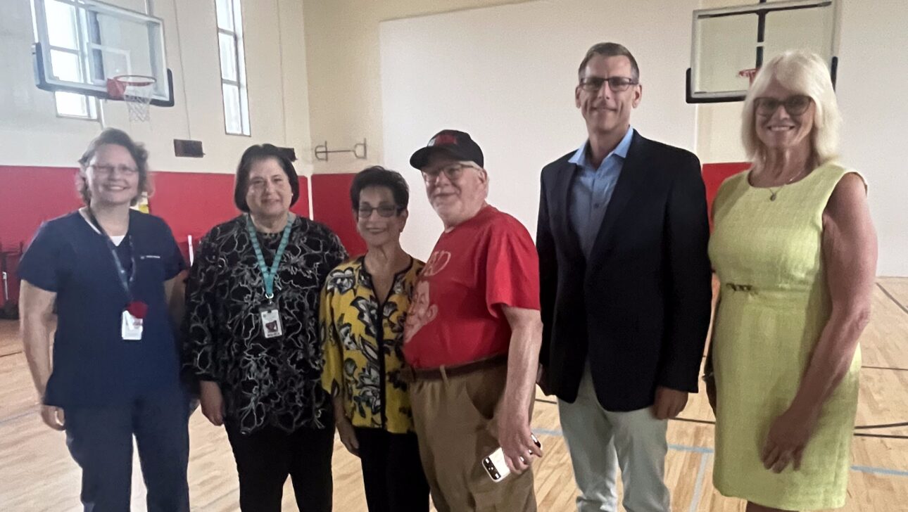 Six adults stand together in a New York gymnasium with basketball hoops and wooden floors, posing for a group photo after an Aging in Community film screening.