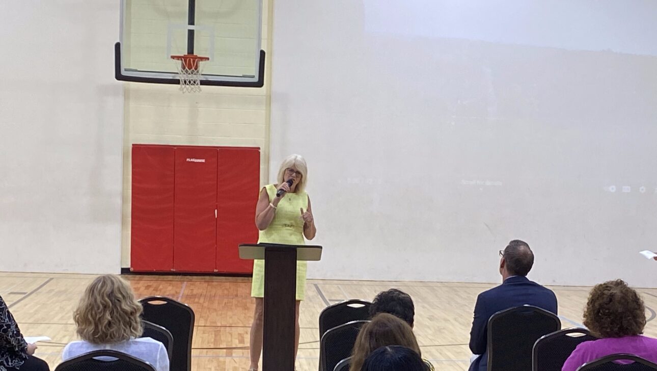 A woman stands at a podium speaking to an audience in a gymnasium in New York, with a basketball hoop and red padding visible in the background during an Aging in Community event.