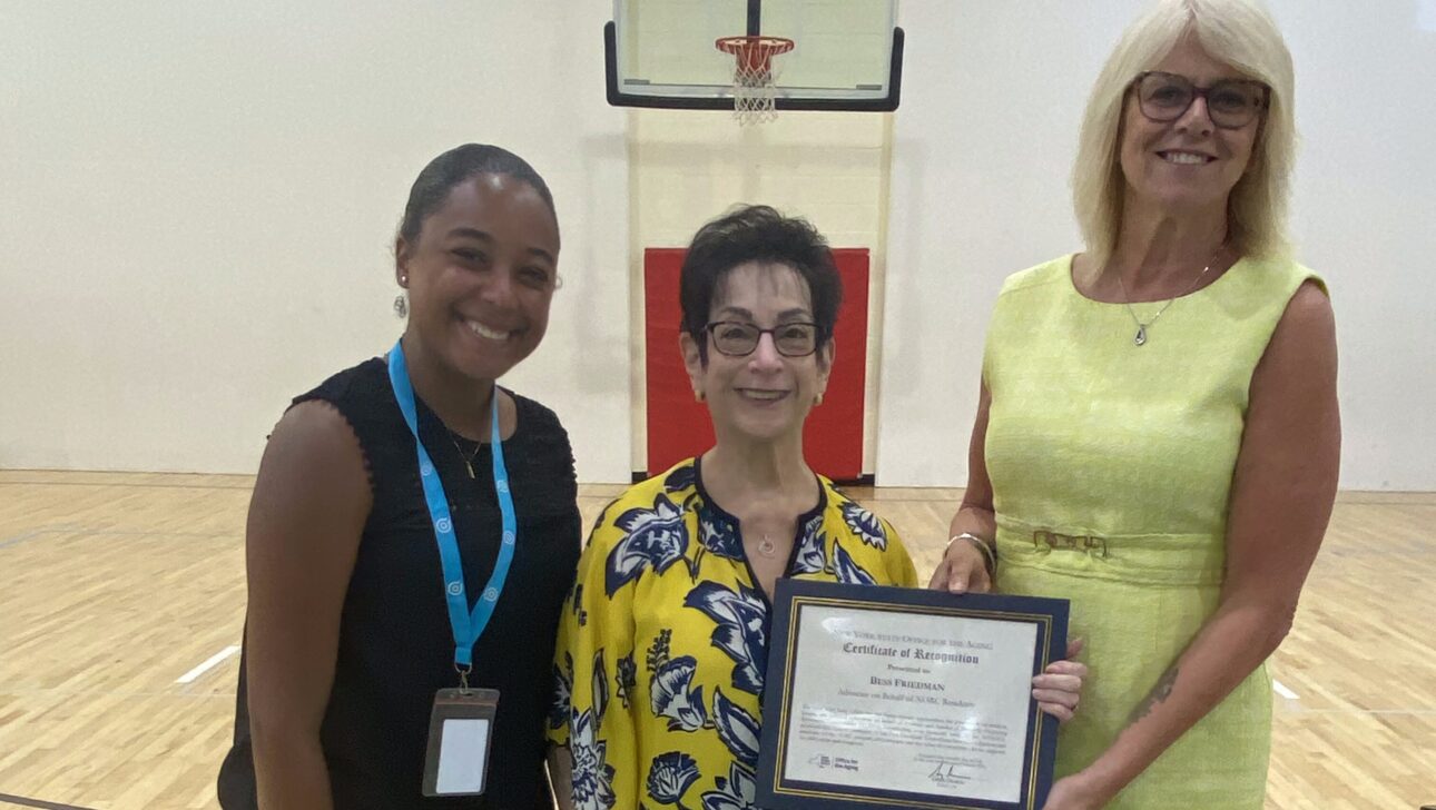 Three women stand on a gym court in New York; one in the center holds a framed certificate of recognition award for her work in Aging in Community.