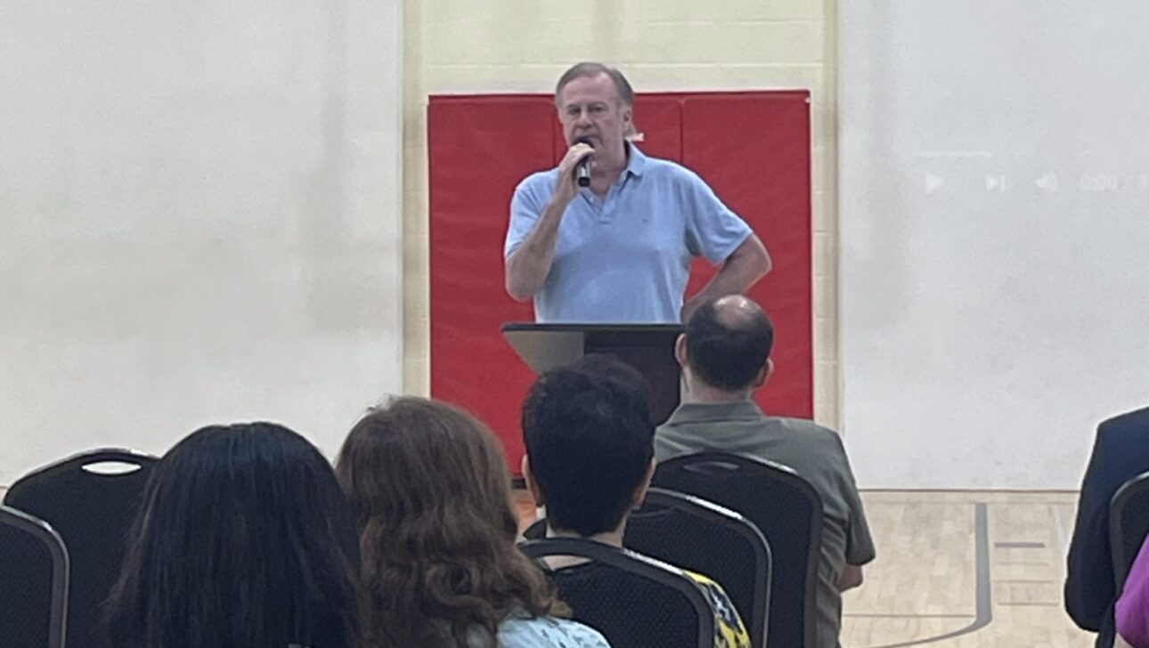 A man stands at a podium speaking to a seated audience in a New York gymnasium, with a basketball hoop visible behind him.