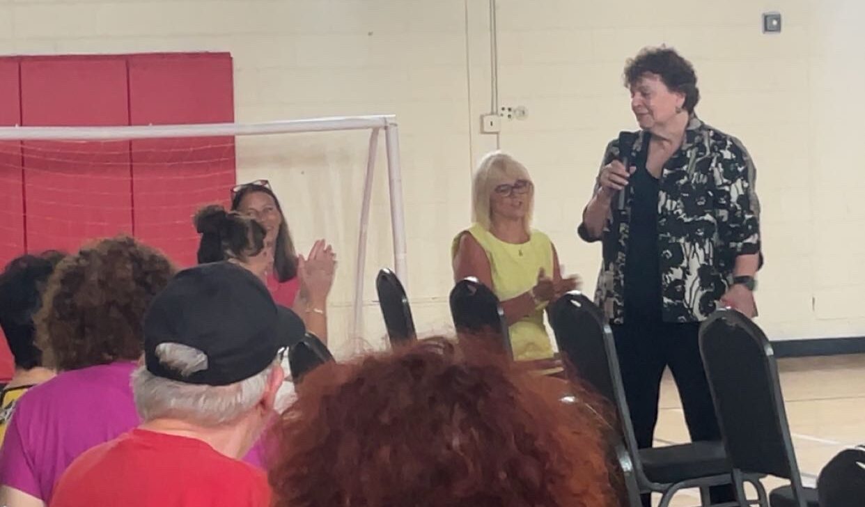 A woman speaks into a microphone in front of a seated audience in a New York gymnasium during an Aging in Community film screening, with a scoreboard and American flag visible in the background.